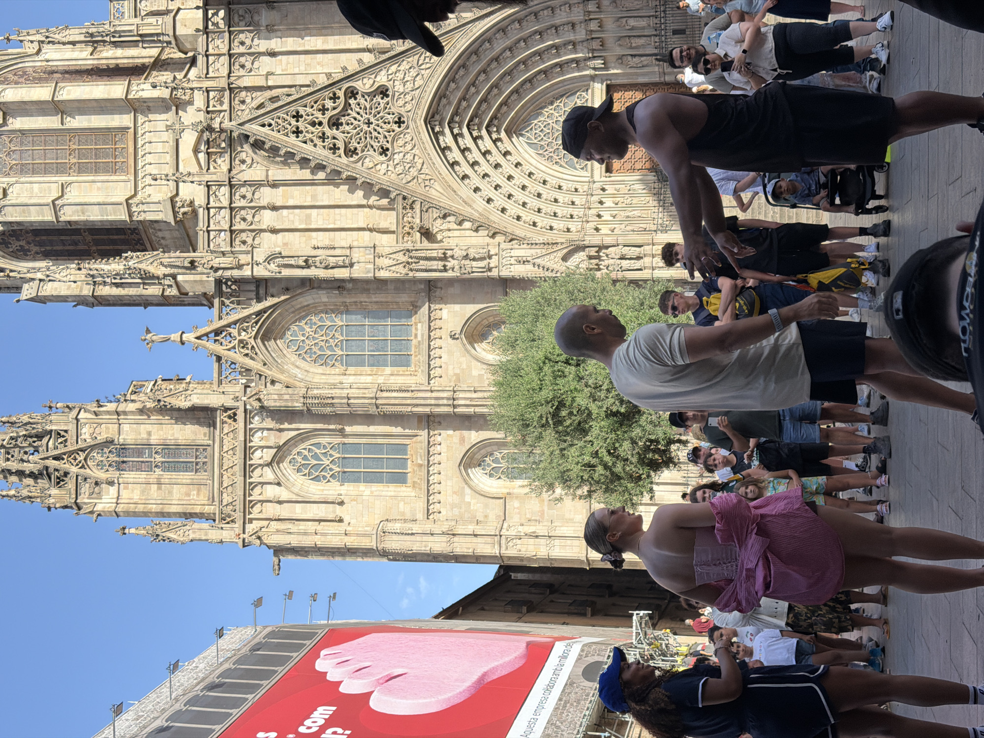 The media file captures a vibrant outdoor scene in Barcelona, Spain, with a prominent Gothic cathedral as the backdrop. A diverse group of people is gathered on a paved area in front of the cathedral's imposing facade, which features intricate stone carvings and arched doorways. The time of day appears to be daytime, with clear blue skies and ample sunlight, suggesting fair weather.
The people in the foreground and midground are casually dressed, with some wearing t-shirts, shorts, and hats, indicating a relaxed atmosphere. One individual is holding a phone, possibly taking photos or videos. The composition suggests a gathering or a tourist activity, with people looking in various directions, some at the cathedral, others interacting with each other.
In the background, more people are visible, and a large red banner with an image of a pink foot and text is displayed on a building. The text on the banner appears to be in Spanish, with visible words like "com" and "Aquesta empresa col·labora amb la millora de".
The overall impression is one of a lively public space within a historic European city, where both locals and tourists are enjoying the architecture and the pleasant weather.