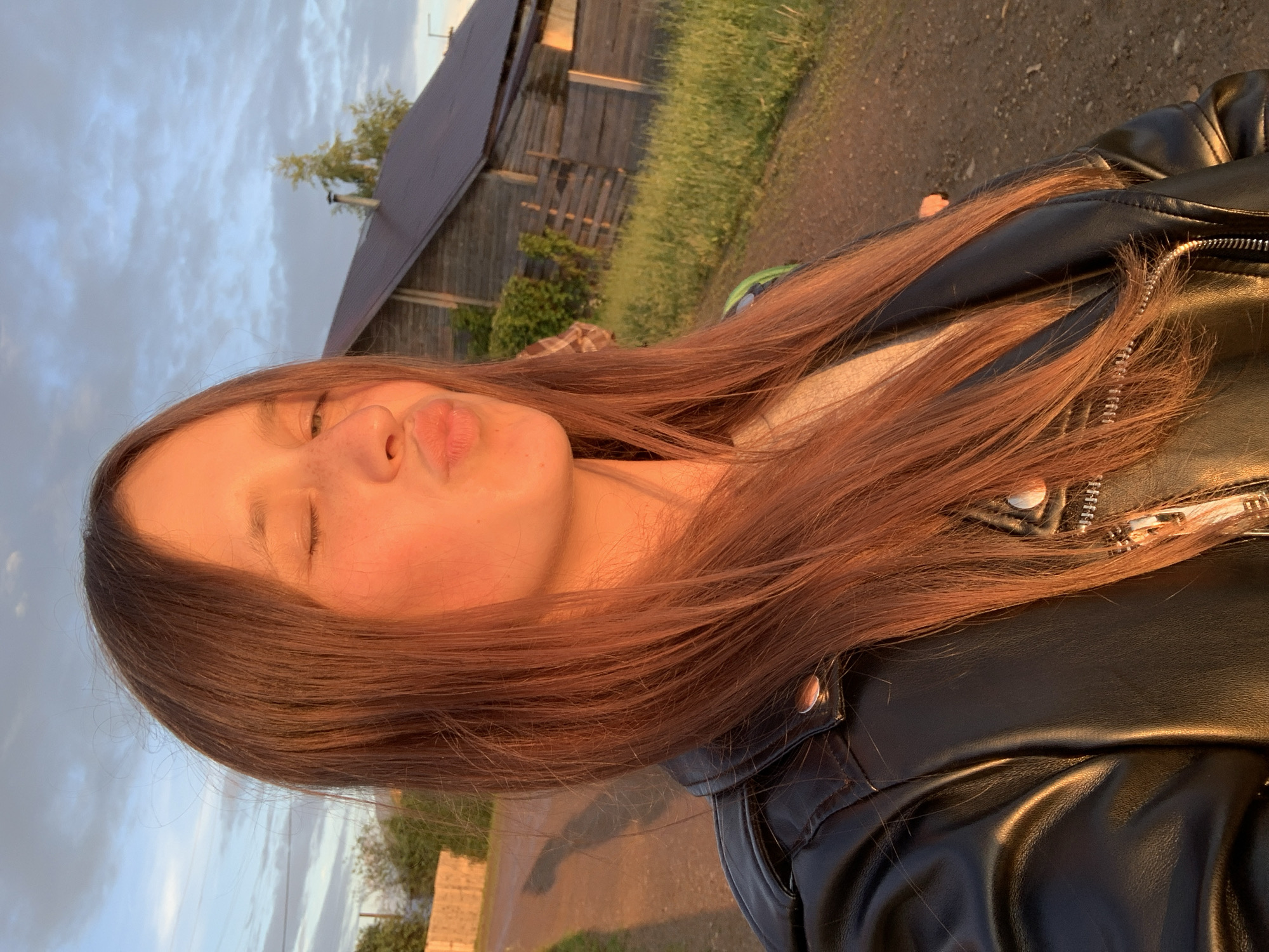 The image is a selfie taken outdoors in a rural setting in Фоки, Russia. The subject is a young person with long, reddish-brown hair, wearing a black leather jacket. Their eyes are closed, and they are puckering their lips as if blowing a kiss or making a playful expression. The warm, golden light suggests it is either sunrise or sunset, with the sun casting a warm glow on the subject's face and hair.
In the background, there is a rustic wooden structure, possibly a barn or an old house, with a dark roof. Adjacent to it, there's a patch of green grass and a dirt path. The sky is partly cloudy with a mix of grey and blue hues. The overall atmosphere is relaxed and casual, conveying a sense of youthful playfulness and enjoying the natural light of a rural evening or morning. There is no visible text in the image.