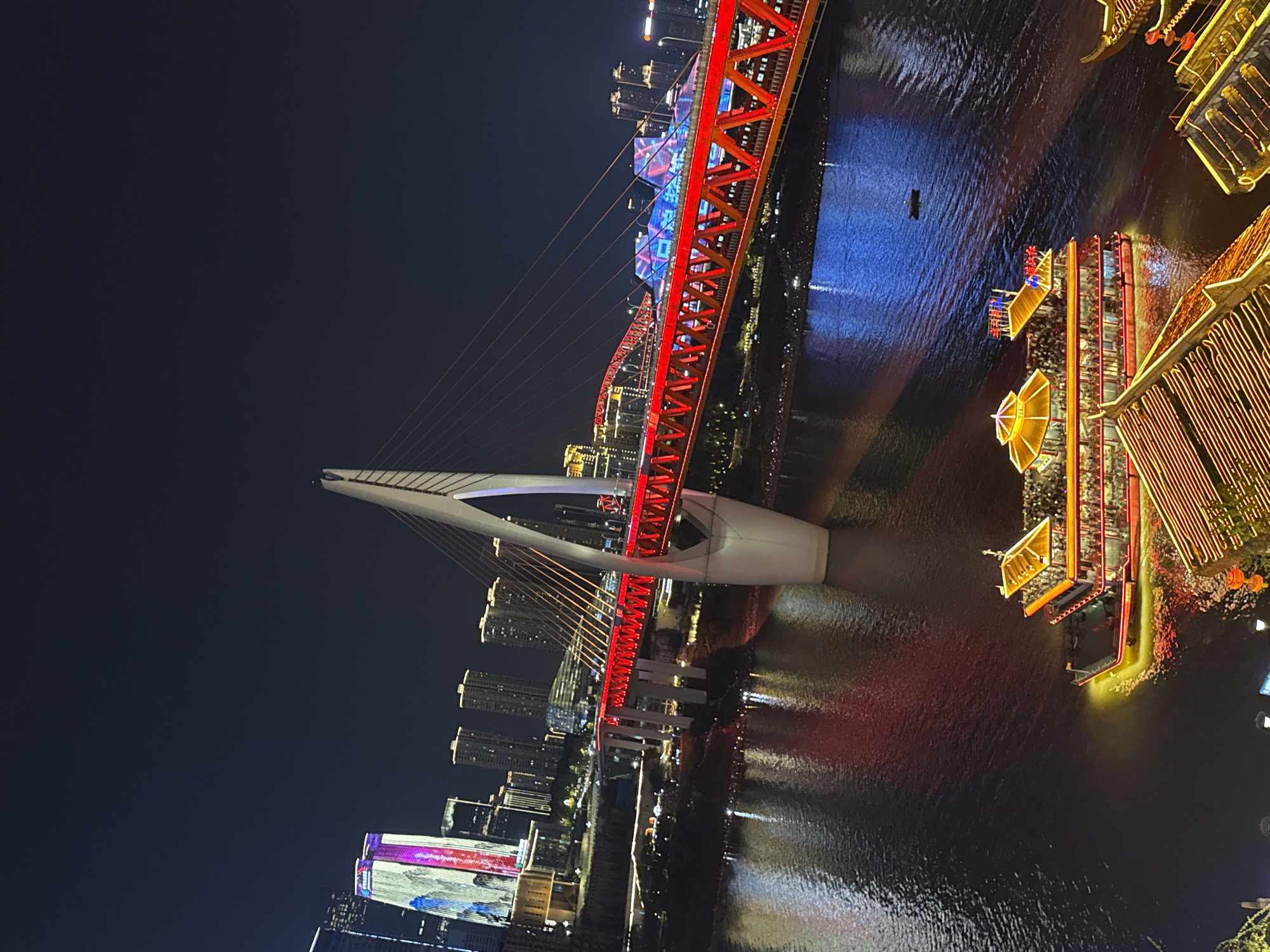 The image captures a vibrant nighttime scene in the Yuzhong District of China, likely along the Yangtze River. A large, illuminated cable-stayed bridge dominates the midground, with its red and blue lighting creating a striking contrast against the dark sky. The bridge's white pylon and suspension cables are clearly visible. Below the bridge, the dark water of the river reflects the colorful lights from the bridge and the surrounding cityscape.
In the foreground, a multi-story tour boat, brightly lit with yellow and red lights, cruises on the river, filled with numerous passengers. The boat has a distinctive tiered roof structure. To the right and bottom of the image, parts of traditional Chinese-style buildings with ornate roofs, also illuminated, are visible, suggesting a riverside promenade or development.
In the background, a cluster of tall skyscrapers forms the city skyline, with some buildings displaying illuminated facades and advertisements. The overall impression is one of a bustling urban environment at night, characterized by impressive architecture, vibrant lighting, and river activity.