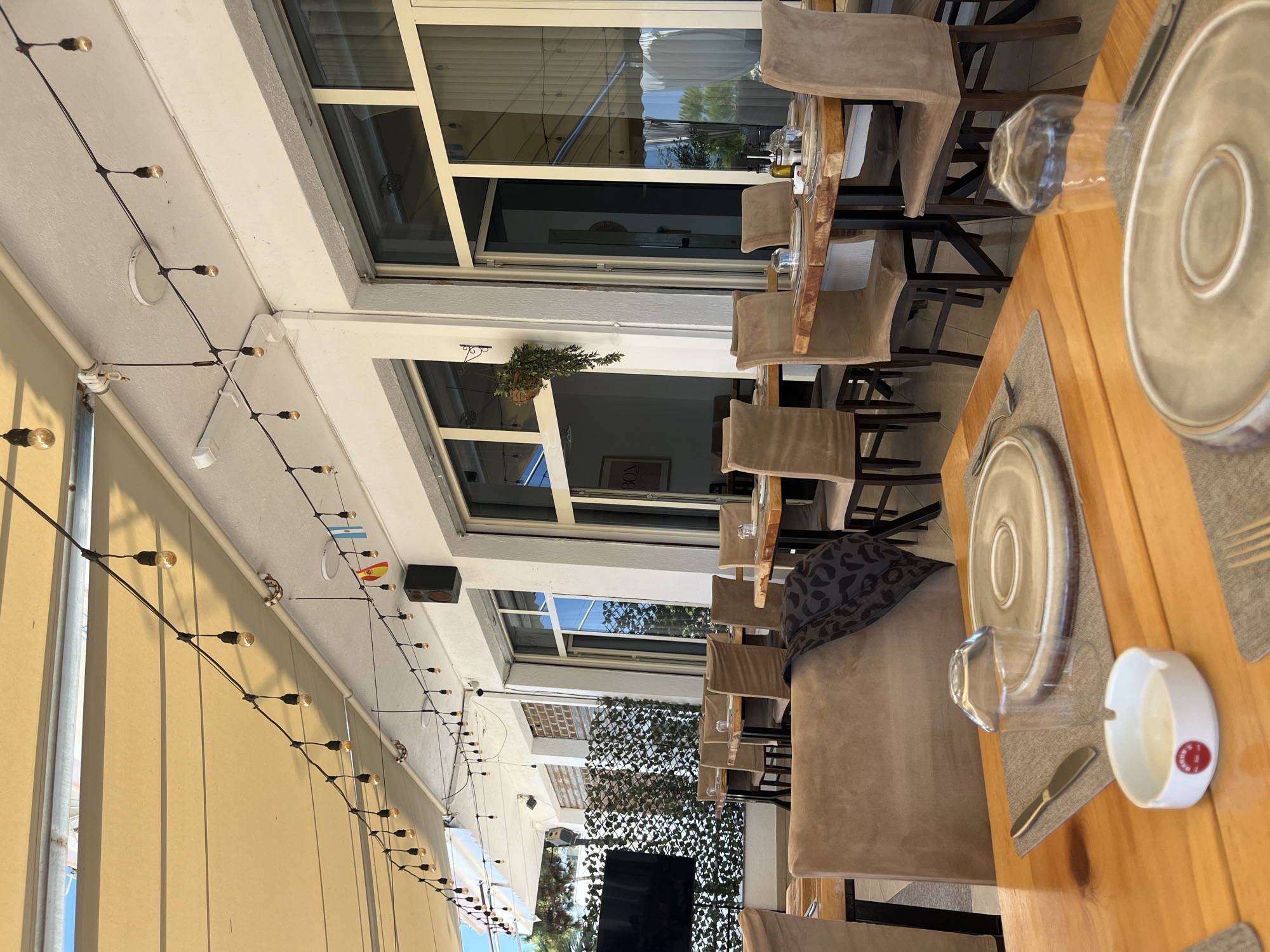 The image shows an outdoor seating area of a restaurant in Bashkia Durrës, Albania, on a sunny day. String lights hang overhead from a beige awning that covers part of the patio. Several tables are set with plates, cutlery, glasses, and napkins. The chairs are upholstered in a light brown fabric. In the background, there are large windows and doors leading into the restaurant. A black speaker is mounted on the wall. A television screen is visible to the left, and a decorative fence or screen is behind it. The overall atmosphere is casual and inviting.