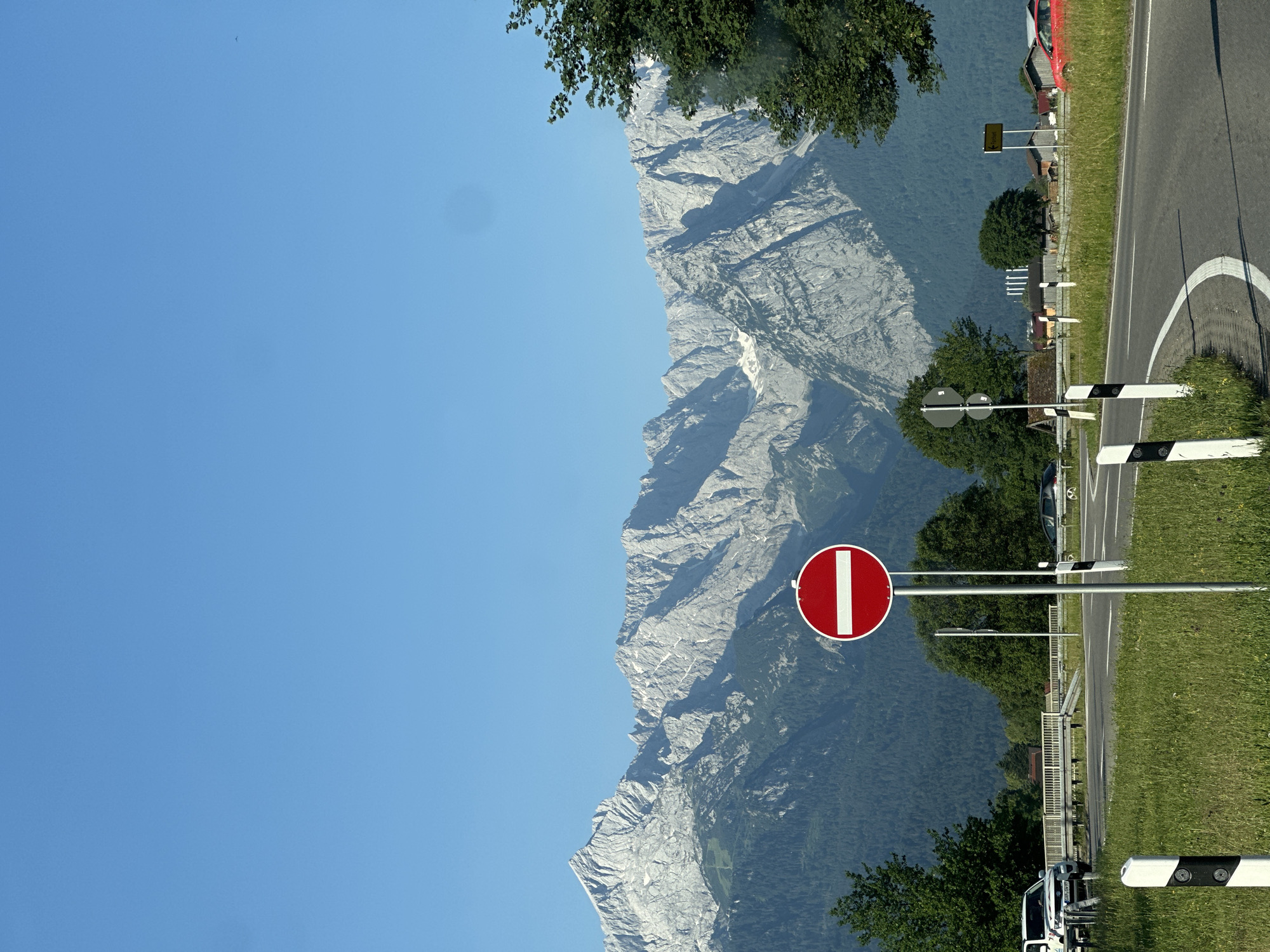 This image depicts a scenic view of the Zugspitze massif, Germany's highest peak, as seen from a road in or near Garmisch-Partenkirchen. The foreground features a winding asphalt road with white lane markings and white-and-black striped bollards on the grassy verge. A prominent red and white "no entry" sign stands on a metal pole. Beyond the road, there is a lush green meadow dotted with wildflowers. In the middle ground, a white SUV is parked on the left, and a red car is visible further down the road on the right. A wooden bridge or overpass spans the road on the left. A cluster of trees and buildings, possibly a small village or farms, are nestled at the base of the mountains. The dominant feature of the background is the majestic Zugspitze, its rugged grey peaks illuminated by sunlight, with patches of snow visible on its higher slopes. The sky is a clear, vibrant blue. The image appears to have been taken on a sunny day, likely in the late morning or early afternoon, given the angle of the light. There are no people visible in the frame.