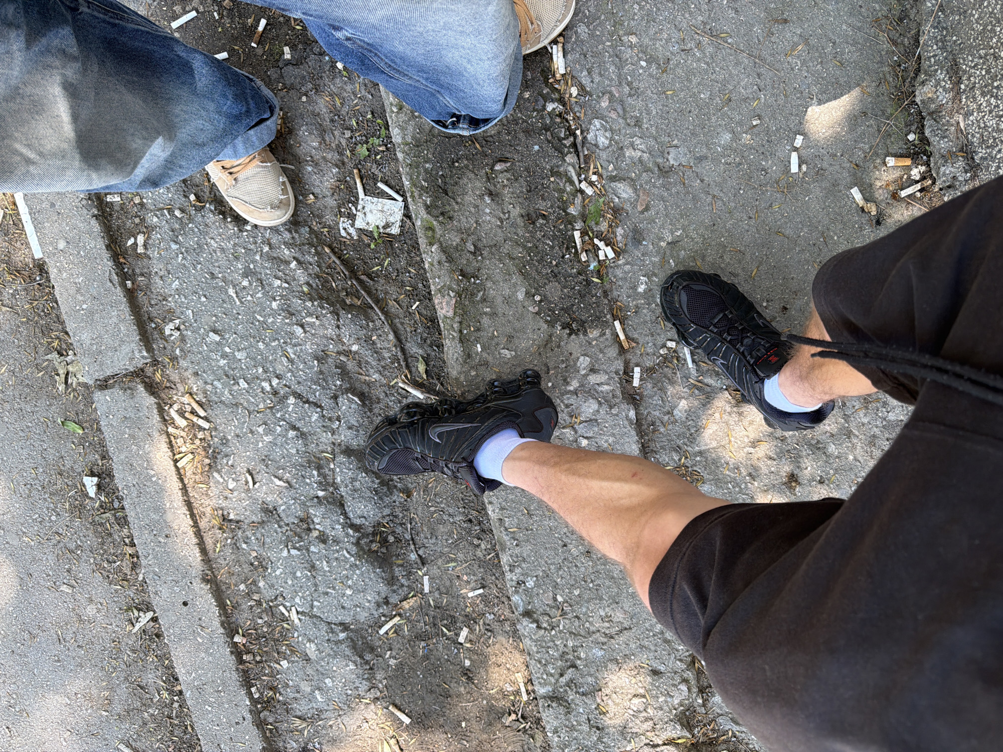 The image depicts a ground-level view of several pairs of legs and feet on a damaged asphalt surface with a concrete curb. The scene appears to be outdoors, possibly in a public space, as evidenced by the accumulation of cigarette butts scattered across the ground.
One person is wearing black shorts and black athletic shoes with white socks. Another person is wearing blue jeans and light-colored canvas shoes. A third person is also wearing blue jeans. The environment is characterized by the rough texture of the asphalt and concrete, with some dried leaves and debris also present. The lighting suggests it is daytime, with shadows indicating the sun is present.
While the image itself does not contain any specific landmarks to pinpoint the exact location within Plovdiv, Bulgaria, the presence of cigarette butts suggests an area where smoking is common, such as a street, park, or public square. The condition of the pavement might indicate an older, less maintained part of the city, or an area with high foot traffic.