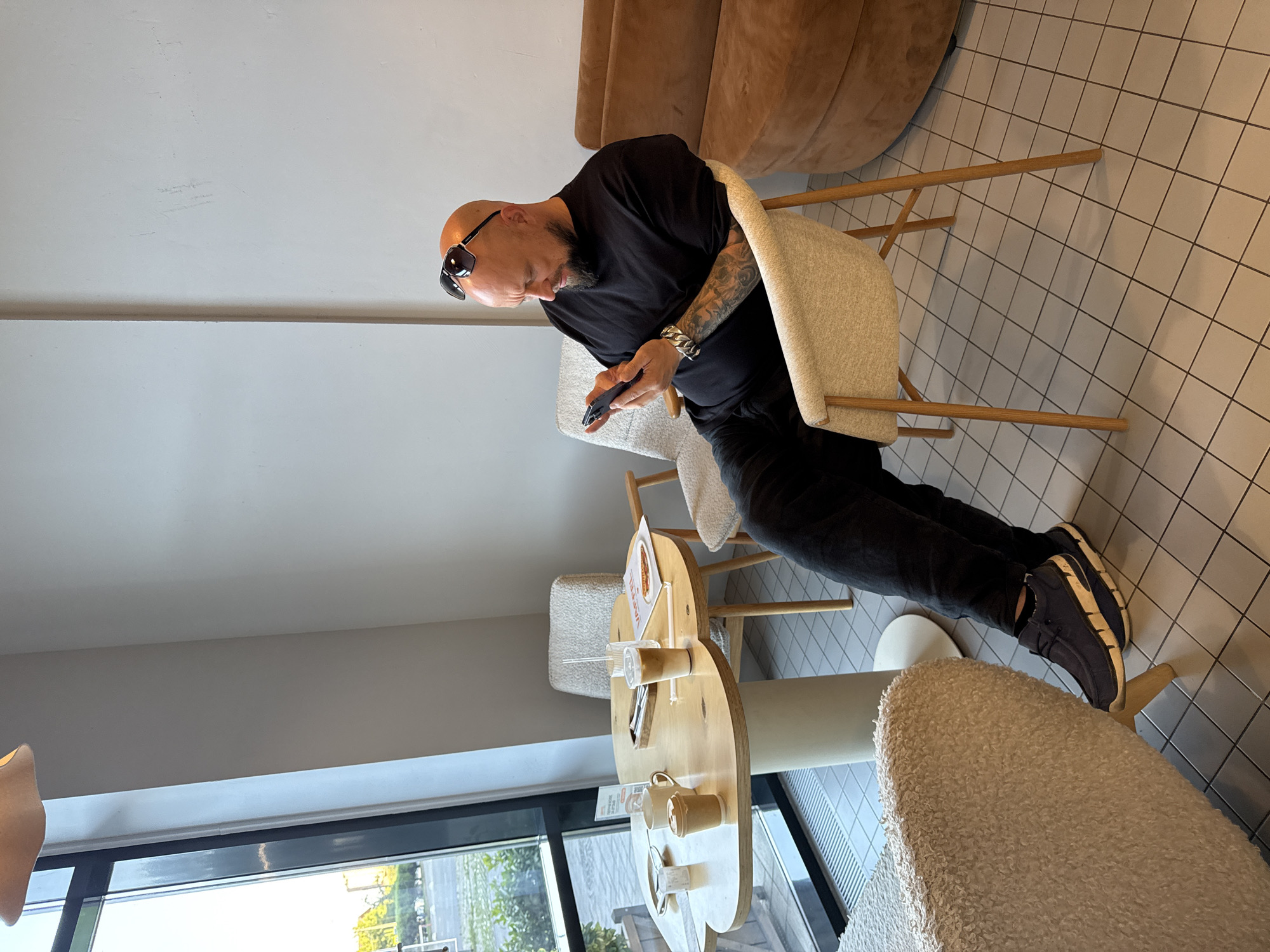 A bald, bearded man with sunglasses on his head is sitting in a cafe, looking at his phone. He is wearing a black t-shirt and black pants, and dark blue shoes. He has tattoos on his left arm and a large metal bracelet on his wrist. He is sitting in a beige chair at a light-colored, cloud-shaped table. On the table are two coffee cups, a glass with a drink, and a menu. In the background, there is a window with a view of greenery, and a brown sofa. The floor is tiled with a grid pattern. The cafe appears to be modern and clean. The image was taken in the city of Krasnodar, Russia.
