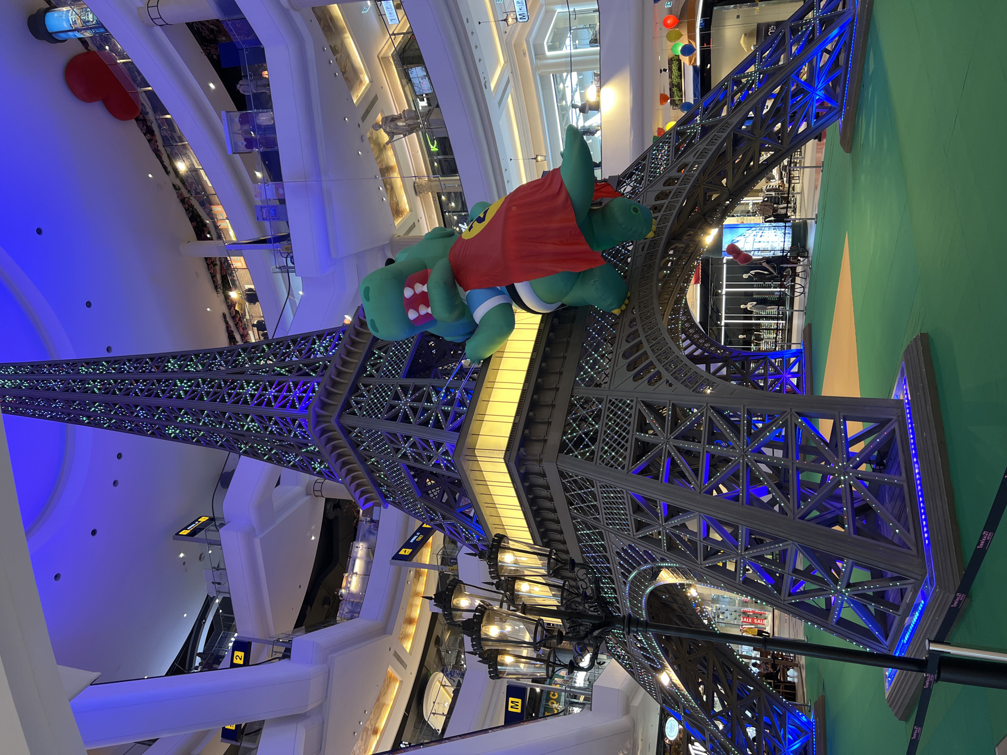 In Pattaya City, Thailand, within the vast, multi-level atrium of Terminal 21 Pattaya, a striking replica of the Eiffel Tower serves as a central display. The tower, constructed with a dark metal framework, is illuminated with numerous green and blue LED lights that accent its intricate lattice design. Atop the tower's arched lower section, a large, inflatable green dinosaur character, dressed in a red cape and a white and blue striped shirt, is positioned as if embracing the structure. The surrounding area is covered in green carpeting, with a tan-colored runway leading into the tower's archway. Several vintage-style street lamps with glowing bulbs stand near the base of the tower, adding to the Parisian theme. In the background, the mall's various levels are visible, lined with shops, escalators, and railings. People can be seen browsing on the upper floors and walking in the distance. Overhead, the mall's expansive ceiling is adorned with soft blue and white lighting, and a large red heart decoration hangs prominently from the ceiling. Colorful balloons are also visible on the right side of the frame, adding a festive atmosphere to the scene.