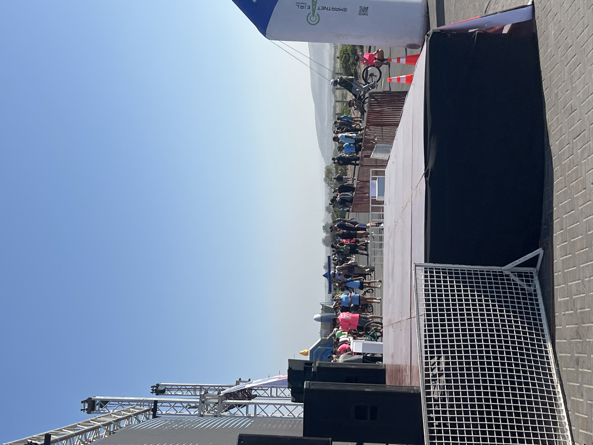 The image depicts a cycling event held outdoors in Arica, Chile, judging by the "SMARTNET EIRL" banner which is a known company located in the city. A line of cyclists, men and women, of mixed ages, are riding on a platform or stage that is elevated. The platform appears to be set up alongside a building or structure, and there are barriers or fences near the edges of the stage. The background shows what appears to be the urban landscape of Arica. The sky is clear and blue. The image seems to have been taken during the event or near its start.