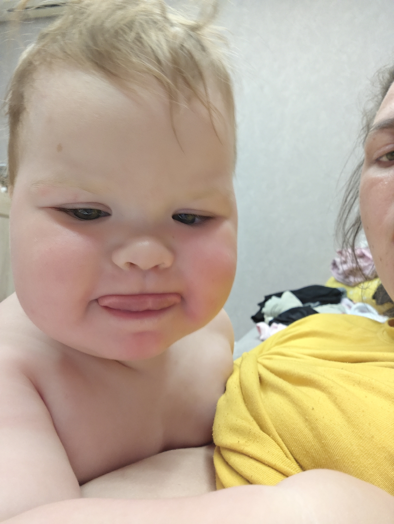 A close-up selfie shows a baby with fair hair and a fair complexion, protruding their tongue in a slightly defiant or playful gesture. The baby is shirtless and appears to be held close to a person wearing a yellow t-shirt. The person is partially visible on the right side of the image, with a portion of their face (an eye, part of their cheek) and upper body in view. In the background, there is a lightly textured white wall and a pile of mixed clothing (black, white, and pink items). The location is unknown and cannot be confirmed from the city of Chaikovsky, Russia, based on this photo alone.