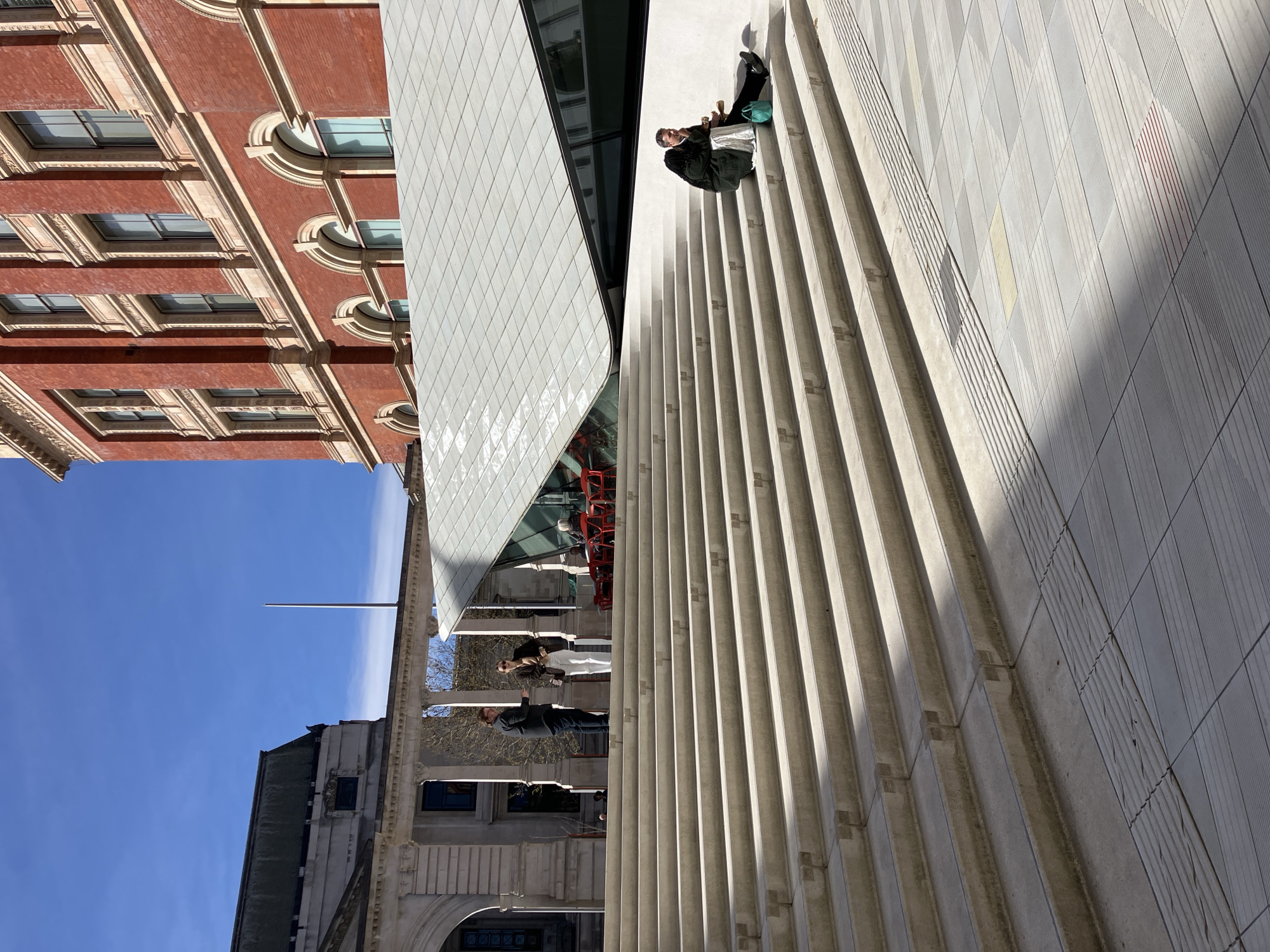 The image depicts the Exhibition Road Quarter of the Victoria and Albert Museum (V&A) in South Kensington, London, United Kingdom. The scene is an outdoor architectural space under a clear blue sky with bright sunlight.
In the foreground, a wide, light-colored concrete staircase ascends diagonally from the lower right to the mid-left. The steps feature small, 'T'-shaped protrusions at their edges. To the right of the staircase, the ground is covered with light grey patterned paving tiles, including tactile paving strips. Sunlight casts distinct shadows across the stairs and pavement.
On the right side of the staircase, a man wearing a dark jacket and dark trousers is seated alone on a lower step. He holds a light-colored object, possibly food, in his hands and looks downwards. A teal-colored bag rests beside him on the step.
Further up the staircase, on a landing, two individuals are standing and conversing. The person on the left is male, dressed in a dark long-sleeved shirt and dark trousers, appearing to gesture. The person on the right is female, wearing a brown jacket over a white top and white trousers, with sunglasses perched on her head. She faces the male.
Behind these individuals and above the staircase is a distinctive modern building with a white, geometrically tiled, sloping roof and angular glass panels. Beneath this structure, an outdoor cafe area is visible, furnished with several red chairs and tables, where indistinct figures are seated.
In the background, to the left, a classical-style stone building with columns is partially visible. To the right, a multi-story Victorian-era building, constructed of red brick with extensive light-colored stone detailing and arched windows, rises above the modern structure. A slender, light-colored pole or spire is discernible against the blue sky in the upper left background.