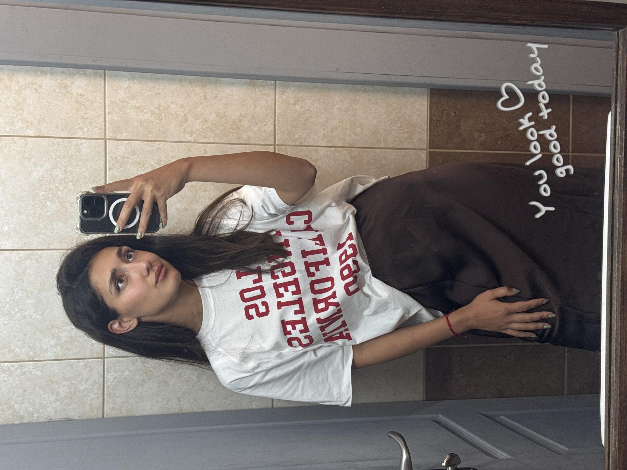 A young adult woman takes a mirror selfie in what appears to be a room with tiled walls, located in Bar, Montenegro. The woman, with long dark brown hair and a tan complexion, looks slightly to her right (viewer's left) with a contemplative expression. She wears a loose-fitting white t-shirt featuring mirrored red text, which reads "LOS ANGELES CALIFORNIA 1989" when un-mirrored. Her lower garment is dark brown, possibly a skirt or wide-leg pants. On her right wrist (left in the reflection), she wears a thin red string bracelet. She holds a dark-colored smartphone, likely an iPhone with a clear case and a MagSafe ring, in her left hand (right in the reflection). Her fingernails are painted a light sage green. The background consists of light beige square wall tiles with darker grout lines, with a row of darker brown tiles at the bottom. A light-colored door with a metallic lever handle is visible to the left (right in the reflection). The mirror itself has a dark brown wooden frame. In the bottom right corner of the mirror, white handwritten text states "You look good today ❤️".