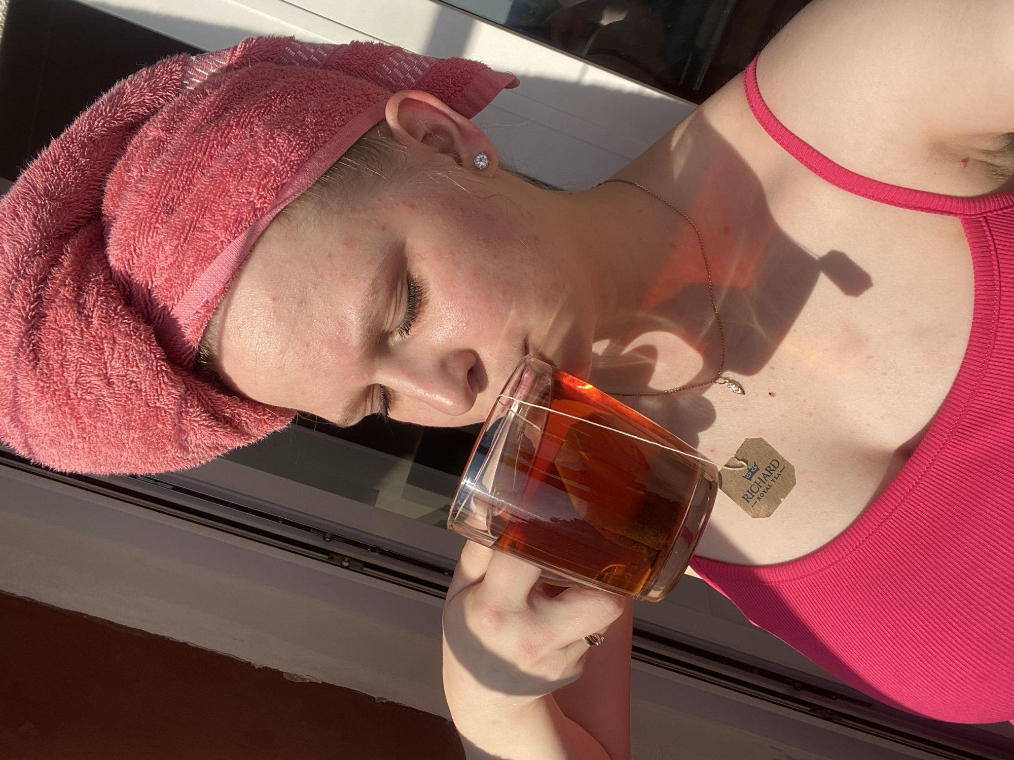 A woman wearing a pink towel on her head and a pink tank top is sitting on a ledge near a window, drinking tea from a clear glass mug. A "Richard Royal Tea" teabag hangs from the mug. She is also wearing a necklace and has a diamond earring. The setting appears to be in Unknown, Unknown.