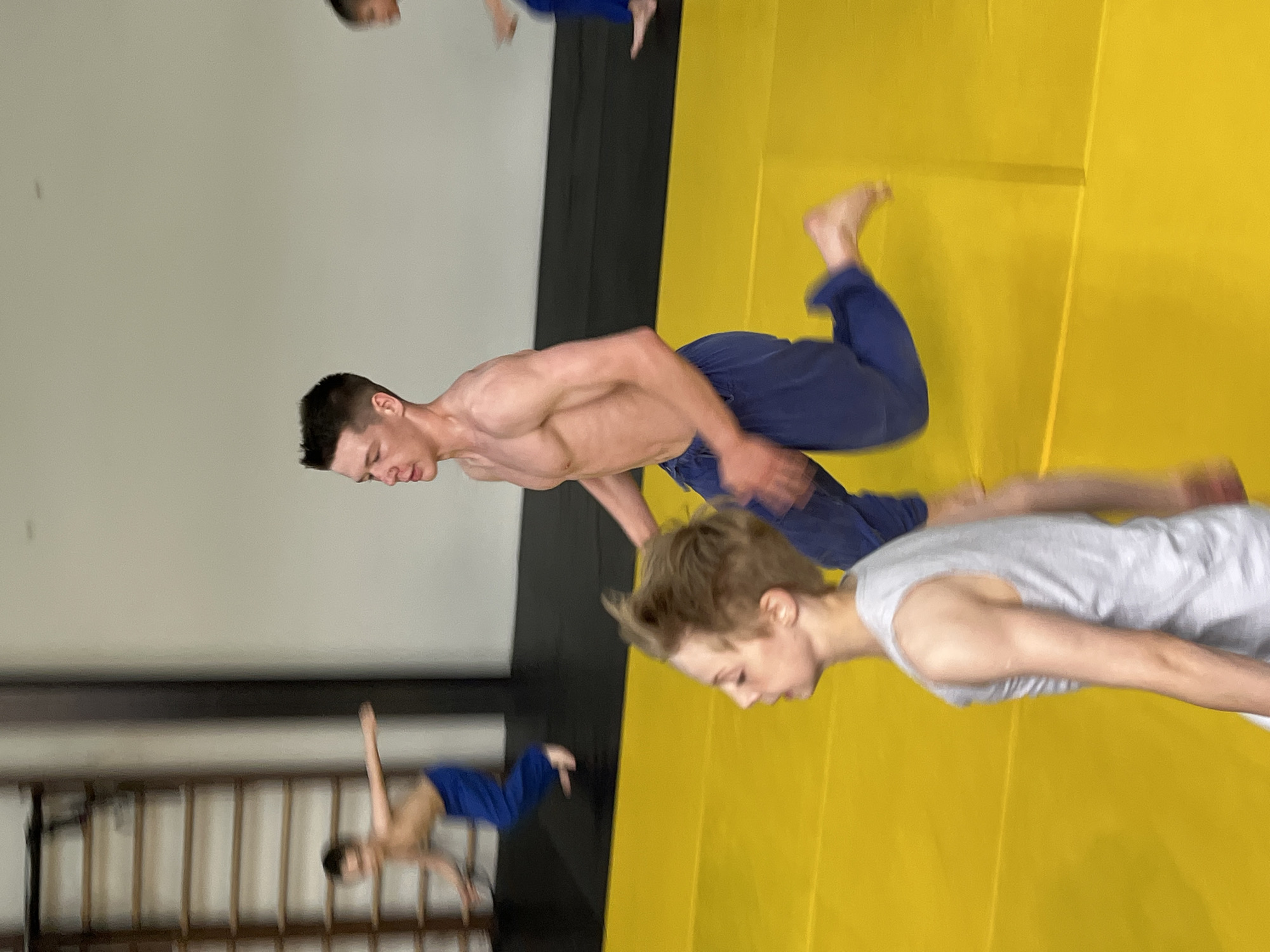 The image depicts an indoor gymnasium with a yellow mat floor. Several young males, shirtless or wearing tank tops and athletic pants, are engaged in physical activity, possibly a training session. One individual in the foreground, wearing a gray tank top, is facing left and appears to be in motion. Behind him, a shirtless young man in blue pants is running, his body angled to the right and his left leg extended. In the background, another individual in blue pants is also running, and a person is partially visible on a wall-mounted ladder. The gymnasium has a neutral-colored wall and a dark mat area along the back. The lighting suggests an indoor environment. There is no visible information indicating the location within the city of Krasnoyarsk, Russia.