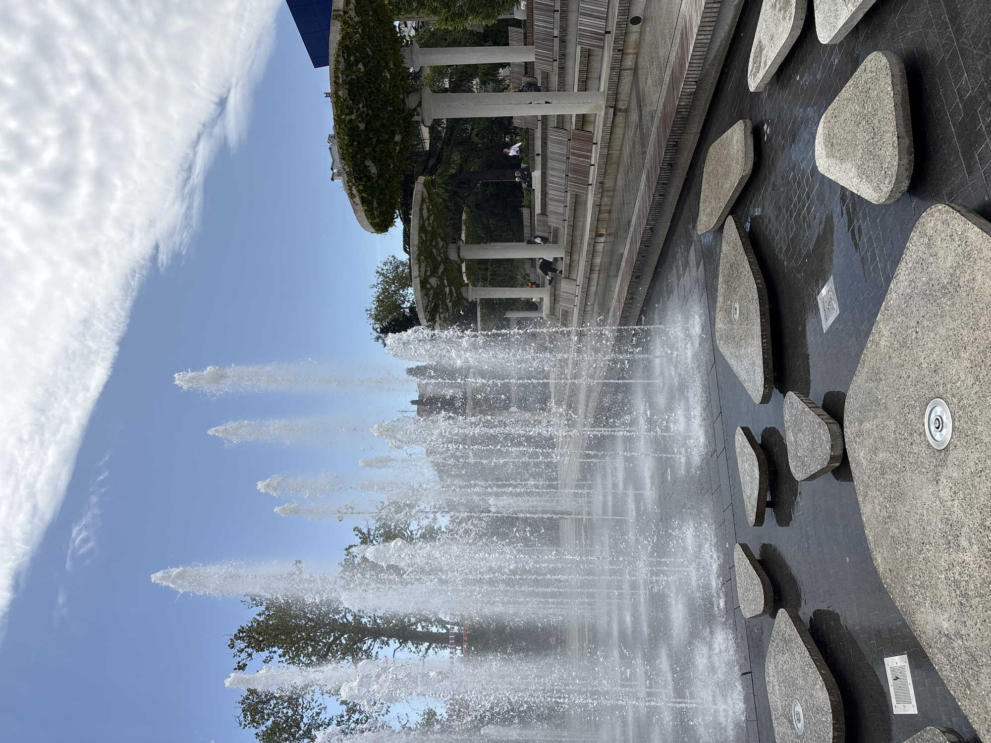 The image shows a fountain with several jets of water shooting upwards into the sky, partially obscuring a view of the city of Yalta, Ukraine. The foreground includes irregularly shaped, smooth gray stones set in a dark, wet surface. Behind the fountain, there's a tiered structure with concrete pillars supporting a pergola covered with greenery. People are sitting on benches along the tiered levels. The sky is visible in the background, partly clear with some clouds. The angle suggests the photo was taken from a low vantage point, looking upwards.