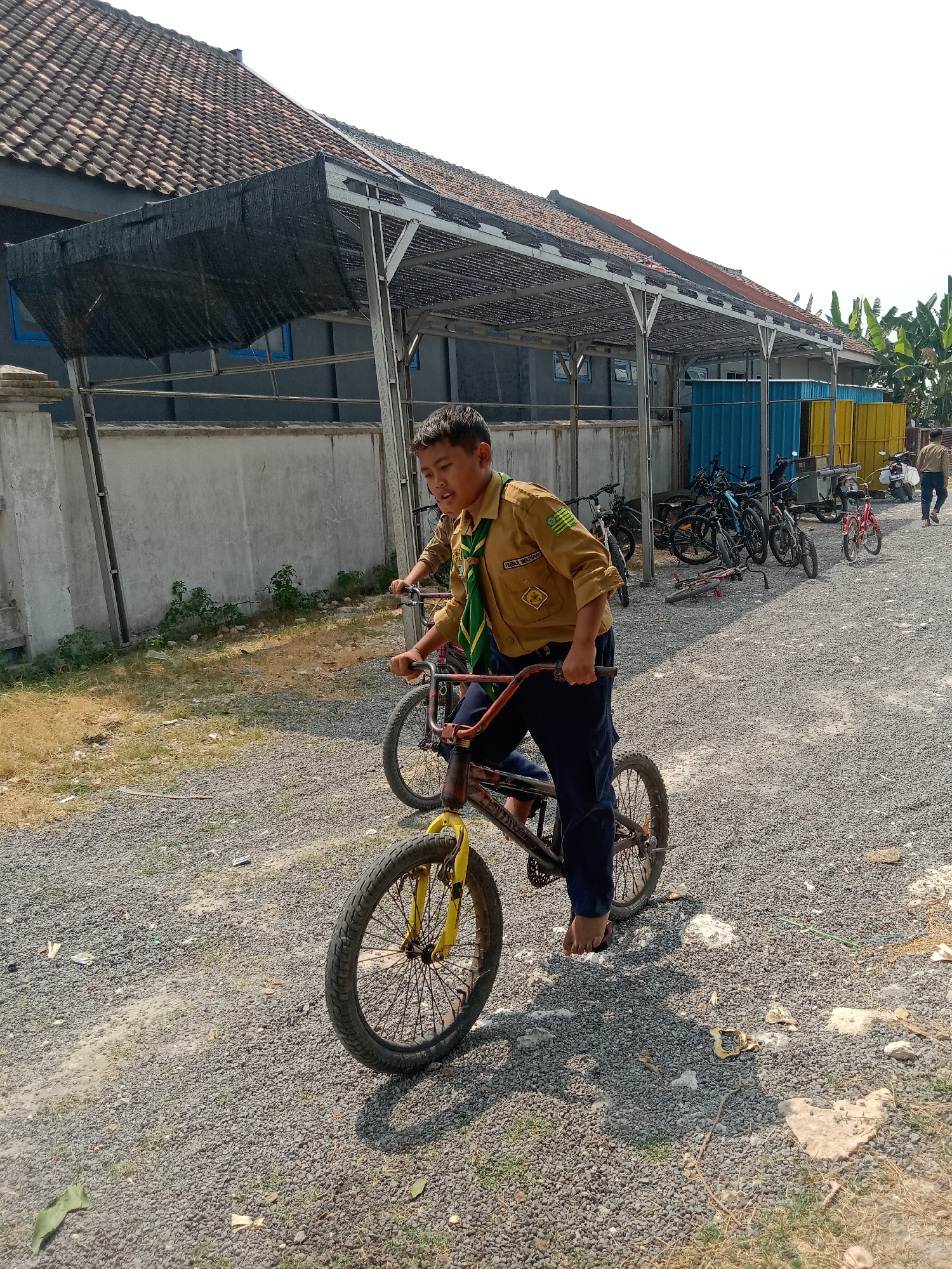 In Sidobandung, Indonesia, on a sunny day, a young boy wearing a tan scout uniform with a green scarf rides a BMX bicycle on a gravel path. He is focused on his ride, his mouth slightly open. Behind him, another person, also in a scout uniform, is visible on a bicycle. The background features a building with a tiled roof and a gray wall, partially covered by a dark mesh awning. Several other bicycles are parked in the distance, and a few people, also in similar uniforms, are seen walking. The environment is dry and dusty, with patches of grass and scattered leaves on the ground. The scene appears to be an outdoor activity area.