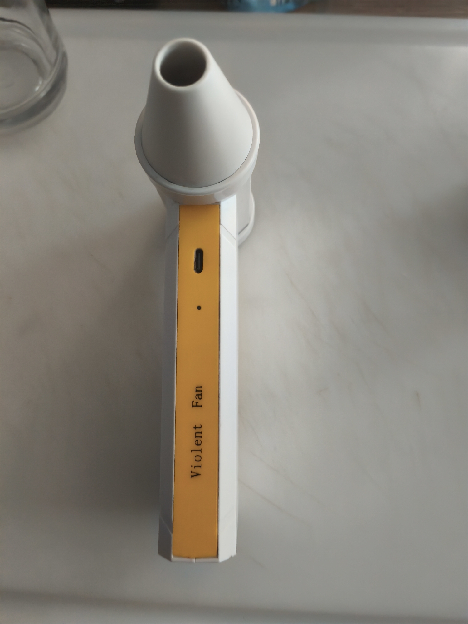 A white and yellow handheld fan with the text "Violent Fan" printed on its side is resting on a light-colored, speckled surface. The fan has a conical white nozzle at the top and a USB-C charging port is visible on the side. A small black dot, possibly a button or indicator light, is also present. In the background to the left, a portion of a clear glass object is visible. The scene is indoors, likely a domestic setting. There is no indication of the city of Assen, Netherlands, or any specific location within it.