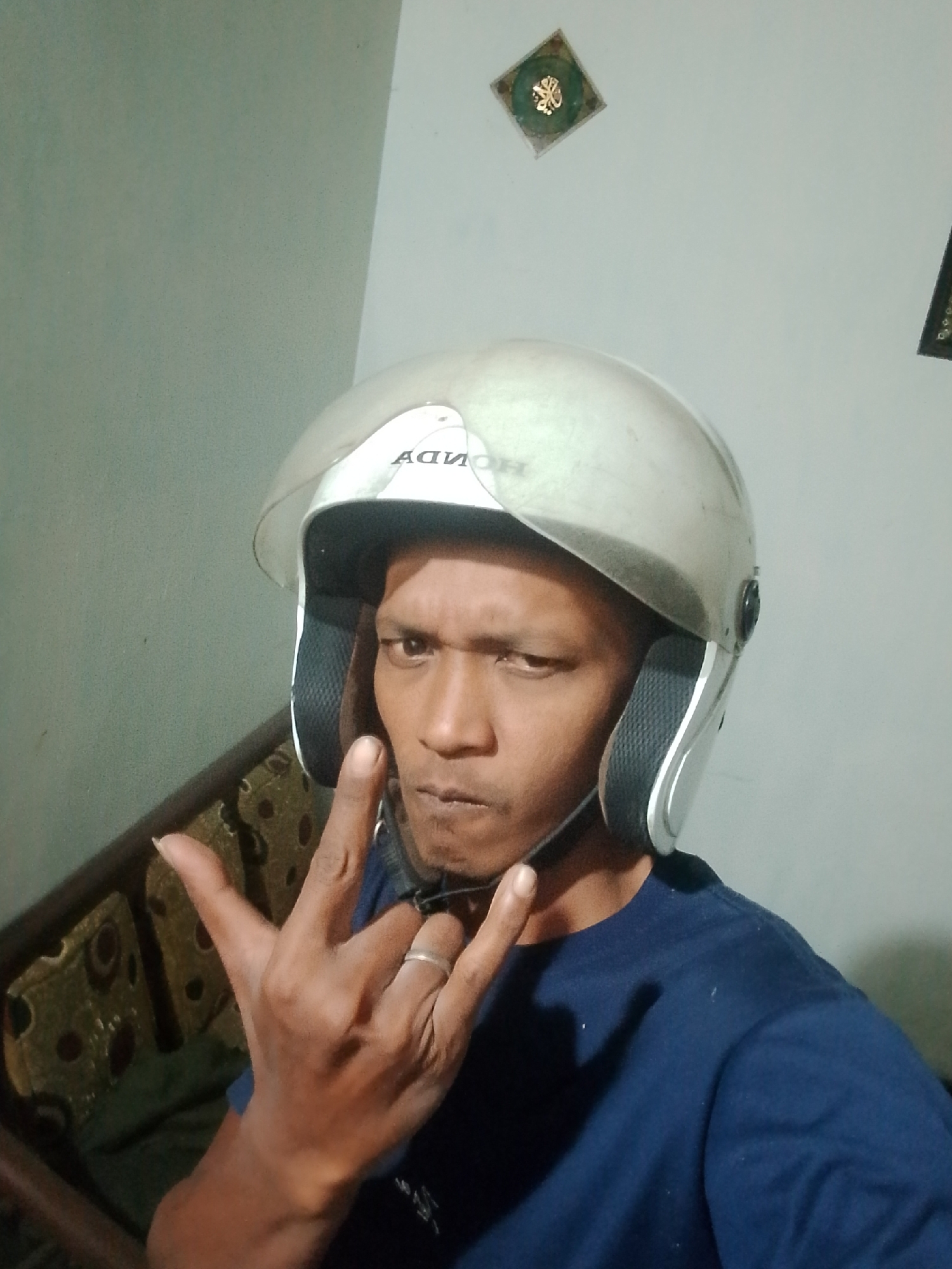A man wearing a silver motorcycle helmet and a dark blue t-shirt is shown from the chest up, looking directly at the camera. He has his left hand raised, forming a "rock on" gesture with his index and pinky fingers extended, and his thumb and middle fingers bent. A silver ring is visible on his ring finger. The man appears to be indoors, with a light green wall behind him and a portion of a sofa or bench with a patterned upholstery visible on the left side of the frame. A small, square decorative item with a green border and gold detailing is mounted on the wall in the upper right quadrant of the image. The lighting suggests it is daytime. The image quality is low, with visible digital noise. The man's facial expression is serious.
