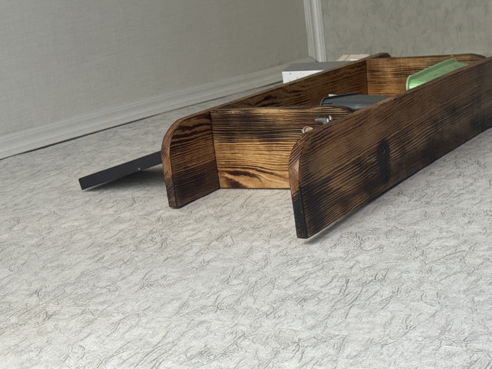 The image shows a dark wooden organizer, likely for a desk or entryway, placed on a textured light-colored surface. The organizer has several compartments and appears to be made from burned wood, giving it a rustic and slightly charred appearance. Inside one compartment, a dark-colored object, possibly a tool or electronic device, is visible. Another compartment contains a small, bright green object, perhaps a piece of stationery or a small accessory. The organizer is positioned at an angle, with its left side slightly lifted by a black metal support. In the background, a light gray wall meets a white baseboard, suggesting an indoor setting, possibly a home or office. The lighting is soft, indicating it might be daytime with diffused light, or an indoor light source. There is no visible text in the image. The scene is static, with no action or event taking place. The overall impression is one of organized storage in a dimly lit environment.