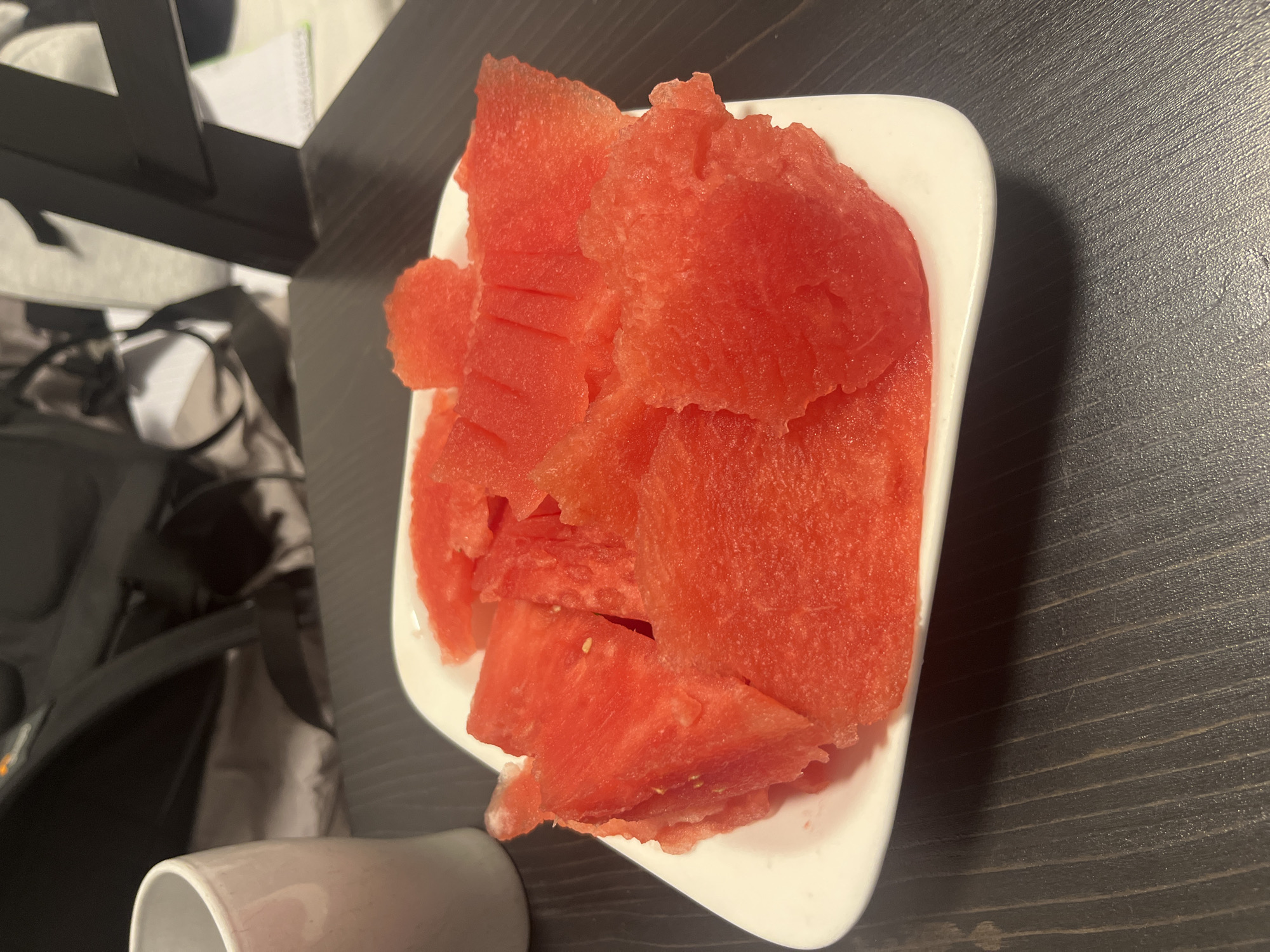 The image displays a white, rectangular dish generously filled with numerous pieces of vibrant red watermelon. The watermelon appears freshly cut, with various irregular shapes and some showing small, pale seeds. The dish is situated on a dark, textured surface, likely a wooden table, which exhibits distinct horizontal grain lines. A portion of a white mug or cup is visible in the bottom left foreground.
The background is out of focus, revealing a somewhat cluttered indoor setting. Visible items include what appears to be part of a black backpack or bag in the mid-left and some light-colored papers. In the upper left, a black, angular object, possibly part of a furniture piece or electronic stand, is also discernible. The lighting suggests an indoor environment, possibly illuminated by artificial light, given the shadows present.
There are no people visible, and no specific activity is taking place beyond the presentation of the fruit. No discernible text is visible on any objects in the image. The scene is set indoors in Tel-Aviv, Israel.