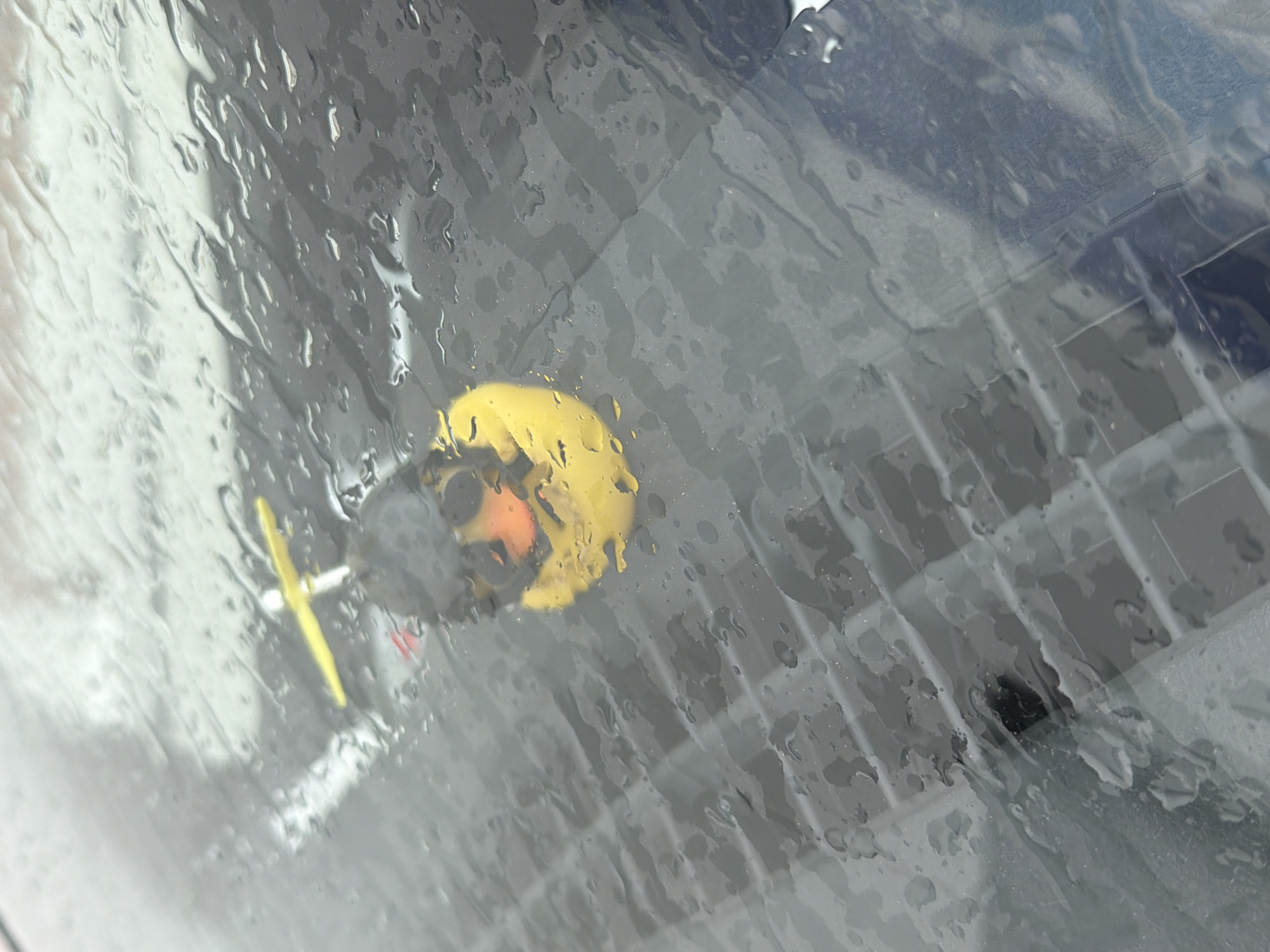 The image appears to be taken through a wet window, possibly a car windshield. Visible through the water droplets are indistinct shapes and colors, including a round yellow object with a smaller dark gray area, and a thin yellow line pointing away from it. The background is blurred, but there appear to be gray and white horizontal lines or bars, possibly metal. It is not possible to determine if the location is Chaikovsky, Russia or identify people or interactions.