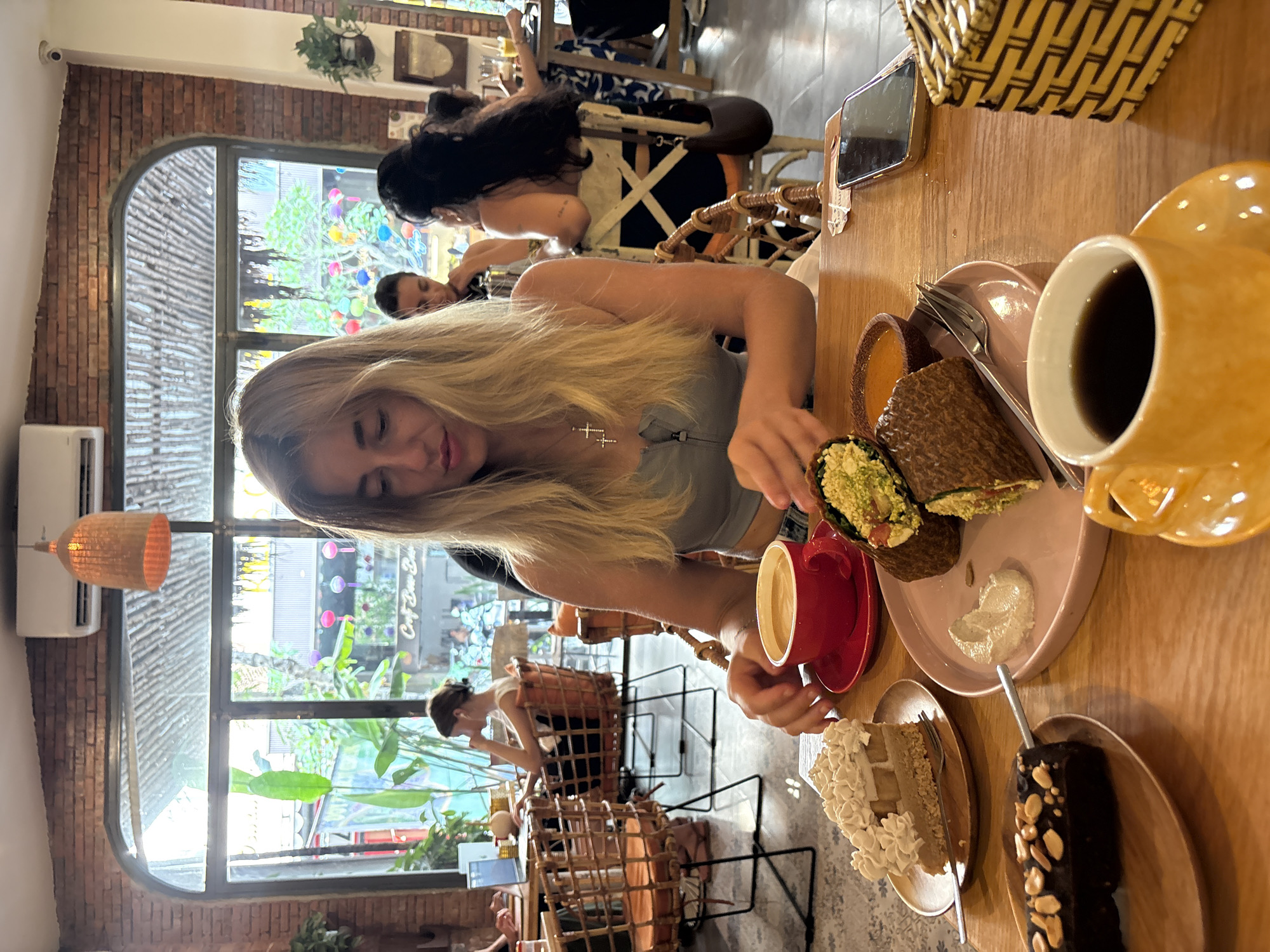 This image captures an indoor scene within a bustling cafe or restaurant, located in Đà Nẵng, Vietnam, during the daytime. The perspective is from a slightly elevated angle, looking across a wooden table.
In the foreground, a young blonde woman is seated, looking down towards her hands and the items on the table. She wears a light-colored top and a prominent cross necklace. Her right hand is holding a red coffee cup, while her left hand is near a large, green-filled wrap on a pink plate.
The wooden table in front of her is laden with food and drinks. Besides the wrap, there's a small bowl of sauce or dip, a yellow/orange mug filled with a dark beverage (likely coffee), and a fork. Further along the table, two slices of cake are visible on separate plates: one is a light-colored cake with a generous amount of white frosting, and the other is a dark, rectangular cake topped with nuts or chocolate chips. A smartphone rests on the table, partially visible.
The background reveals more of the cafe's interior and the outside world through large windows. The cafe features a rustic aesthetic with exposed brick walls. Through the windows, vibrant greenery and hints of other buildings or establishments can be seen. On one of the windows, partially obscured text reads "Cafe Bún Bò," indicating a Vietnamese cafe. Inside, an air conditioning unit and a wicker-style hanging light fixture are mounted on the wall.
Several other patrons are scattered throughout the cafe. One woman with dark hair is visible from behind in the mid-ground, seated at a wicker chair. Further back, another person is seen through the window, and multiple wicker chairs and tables suggest a busy environment. The overall atmosphere appears relaxed and comfortable, with natural light flooding the space.