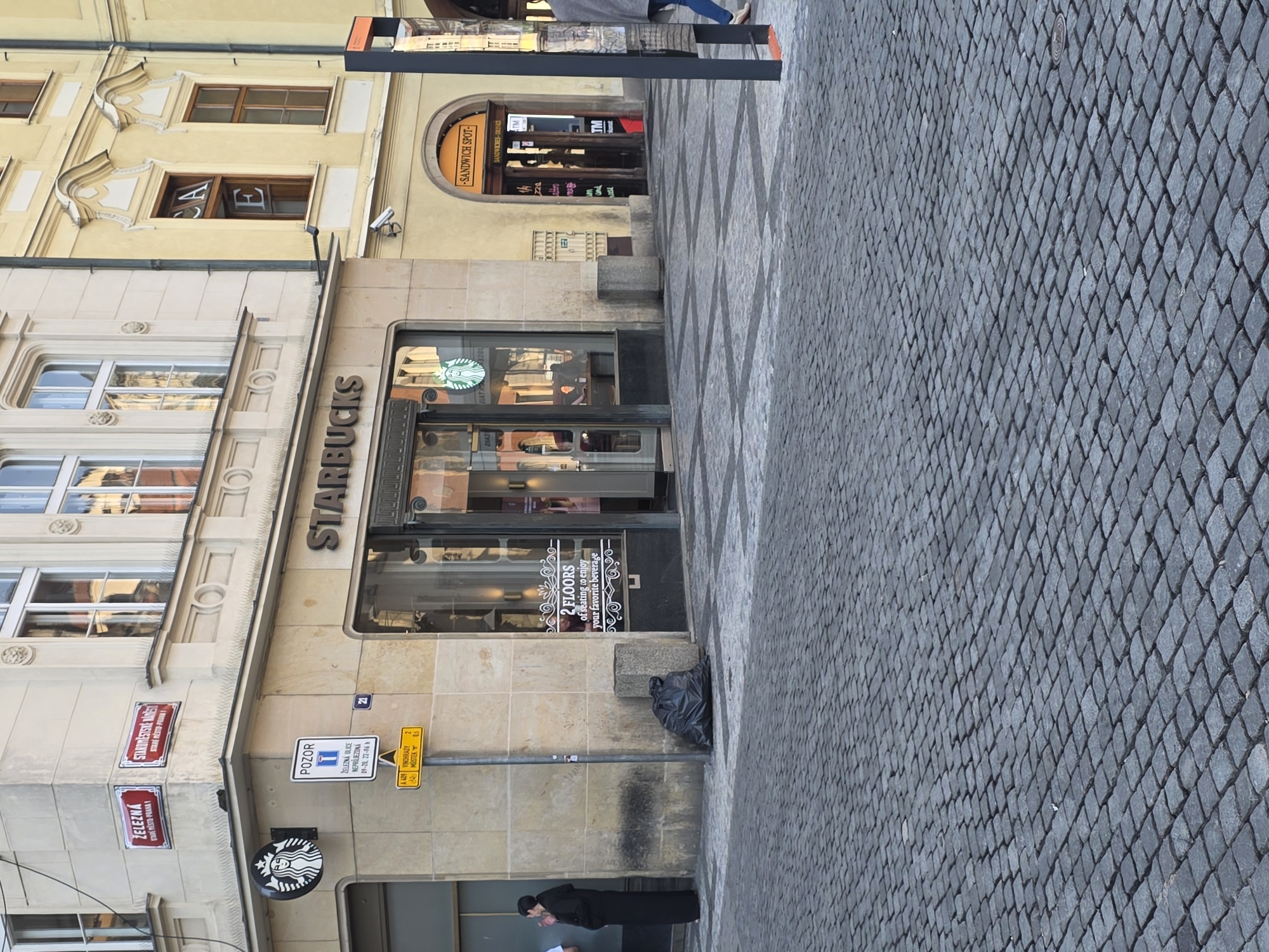 This media file captures a vibrant street scene in what appears to be a historic European city, specifically Prague, Czech Republic, indicated by the Czech street signs. The perspective is looking along a cobblestone street towards several buildings.
The main subject is a multi-story building featuring a Starbucks coffee shop on the ground floor. The Starbucks entrance is prominent, with its name "STARBUCKS" spelled out vertically on the building facade and a circular logo hanging below. A sign in the window advertises "2 FLOORS of seating to enjoy your favorite beverage." Adjacent to the Starbucks, an arched doorway leads to a "SANDWICH SPOT" with a sign that also reads "SANDWICHES - DRINKS." The buildings exhibit classical architectural elements, such as ornate window frames and decorative molding.
The street is paved with gray cobblestones, arranged in a diamond pattern, and features darker contrasting patterns. Several street signs are visible, including red signs for "ŽELEZNÁ" (Železná Street) and "STAROMĚSTSKÉ NÁMĚSTÍ" (Old Town Square). A white sign with a blue symbol reads "POZOR" (Caution) and states "ŽELEZNÁ ULICE NEPRŮJEZDNÁ" (Železná Street Impassable) with specific times "09-00 - 22-00." A yellow directional sign points to "VINOHRADY" and "MÍSTEK."
In the foreground, a person in a dark coat or dress is partially visible near the bottom left, while another person's lower body, wearing jeans and light shoes, walks away in the upper right. A black trash bag rests against the wall near the Starbucks entrance. The time of day appears to be daytime, with clear lighting suggesting fair weather. Reflections of letters "C", "A", and "E" are visible in an upper window of one of the buildings.