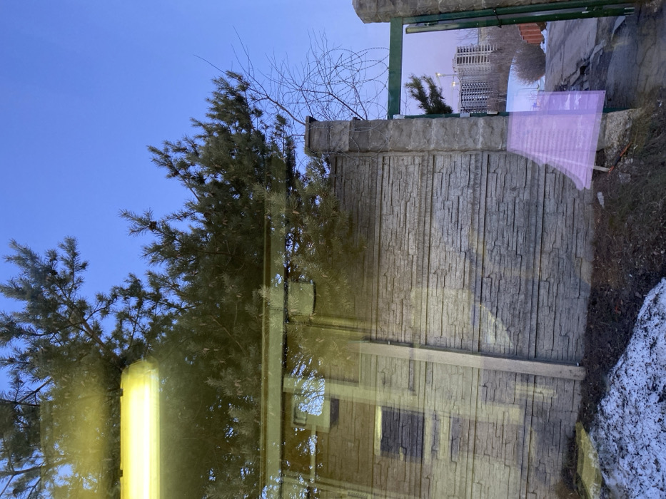 The image depicts an outdoor scene in Марфино, Russia, captured through a reflective surface, likely a window. The upper frame is dominated by a clear, pale blue sky and the dense green needles of a pine tree, with smaller bare tree branches visible to the right. A prominent, horizontal yellow-orange fluorescent light fixture is reflected across the upper-left quadrant.
The mid-ground features a textured, light gray concrete block wall, horizontally patterned to resemble stacked stone. To the right, an open dark green metal gate is visible, flanked by concrete pillars. Coiled barbed wire is present on top of the concrete pillar immediately to the left of the gate opening. Beyond the open gate, a dirt or unpaved road curves upwards, and in the distant background, multiple tall, modern multi-story buildings under construction, including a visible construction crane, stand against the sky.
The foreground shows dark earth with patches of melting snow. A vertical light-colored beam, potentially a wooden post, is positioned near the left side of the concrete wall. A translucent, angular magenta/purple shape is superimposed over the lower-right section of the wall and ground, consistent with a reflection or optical anomaly. No individuals are present in the scene.