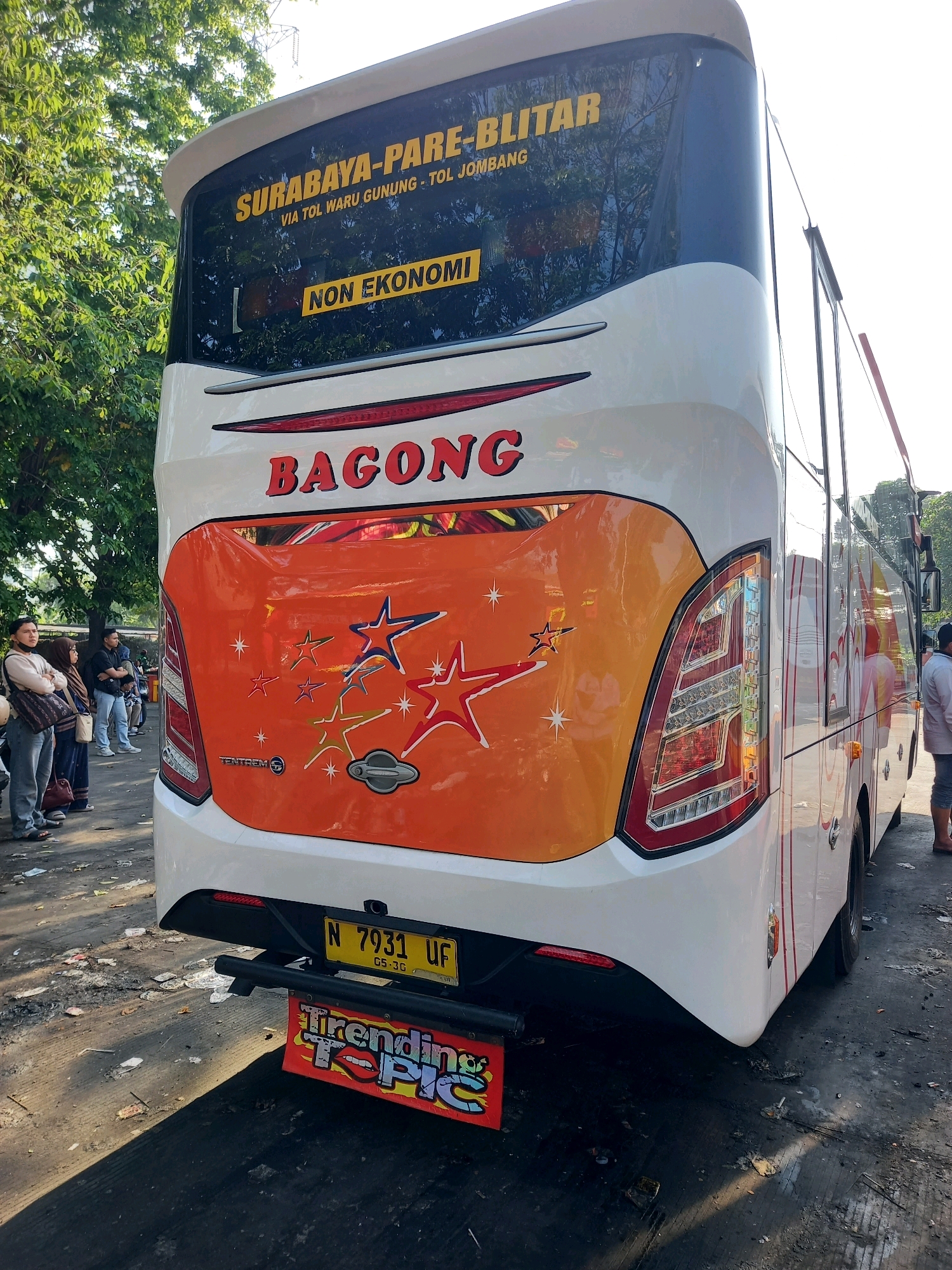 The image shows the rear of a white bus with an orange graphic design featuring stars. The bus has yellow lettering on its rear window that reads "SURABAYA-PARE-BLITAR VIA TOL WARU GUNUNG - TOL JOMBANG" and below that, "NON EKONOMI". The name "BAGONG" is written in large red letters on the back of the bus. Below the bus, a license plate reads "N 7931 UF" and a colorful sign below the bumper says "Trending Topic". The bus is parked on a dirty asphalt surface, possibly in a bus station or parking lot, with trees and some debris visible in the background. Several people are standing near the bus. The overall scene suggests a location in Bungurasih, Indonesia, a well-known intercity bus terminal.