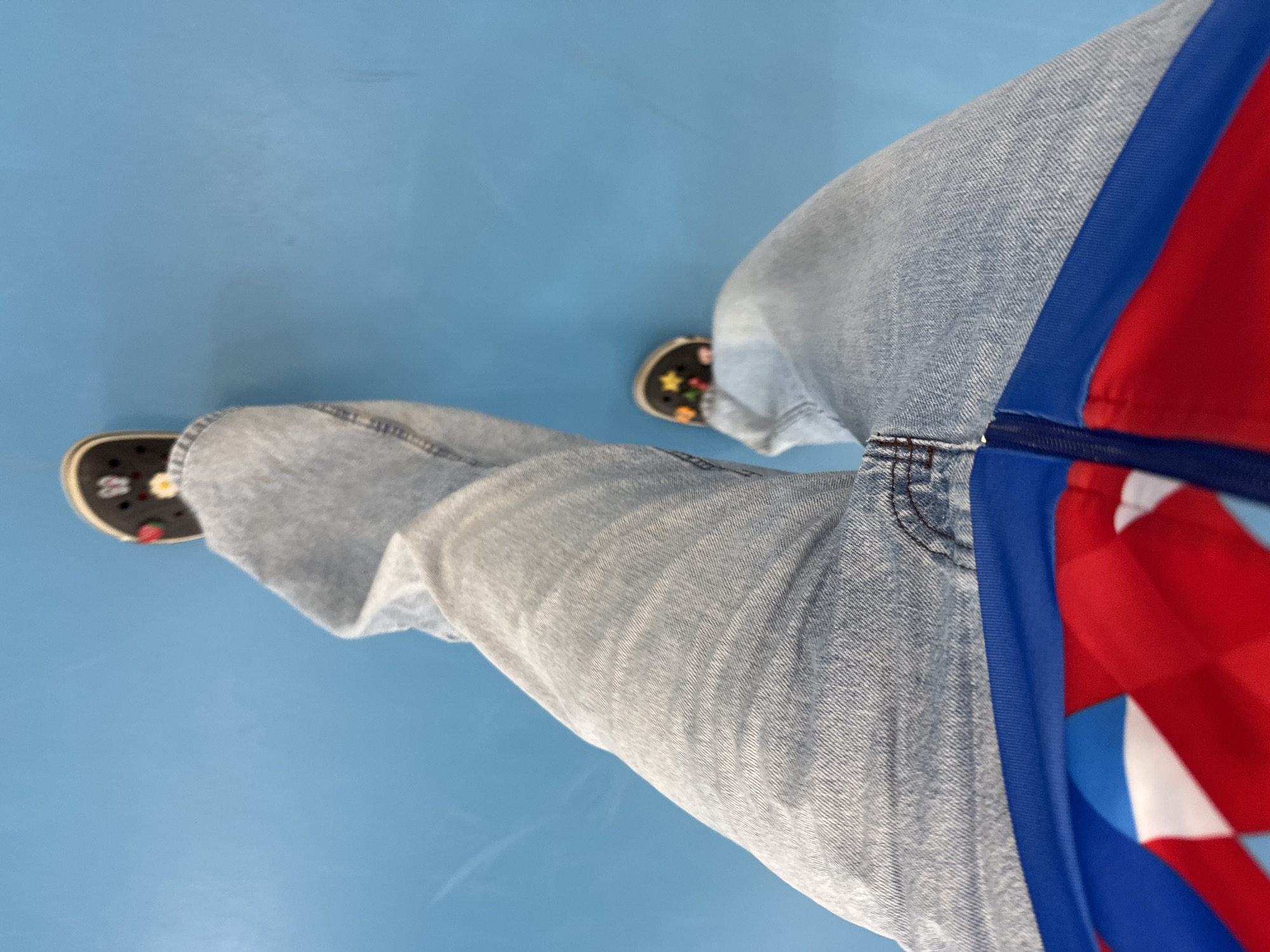 The image presents a downward-angled view of a person's legs and feet positioned on a light blue surface. The individual is wearing light-wash denim pants, adorned with a blue stripe and sections of red and white fabric on the right leg. Their footwear consists of black Crocs-style shoes decorated with colorful, cartoon-like embellishments. The environment appears to be a smooth, solid-colored floor. There is no information to suggest the location is in Usolye, Russia.