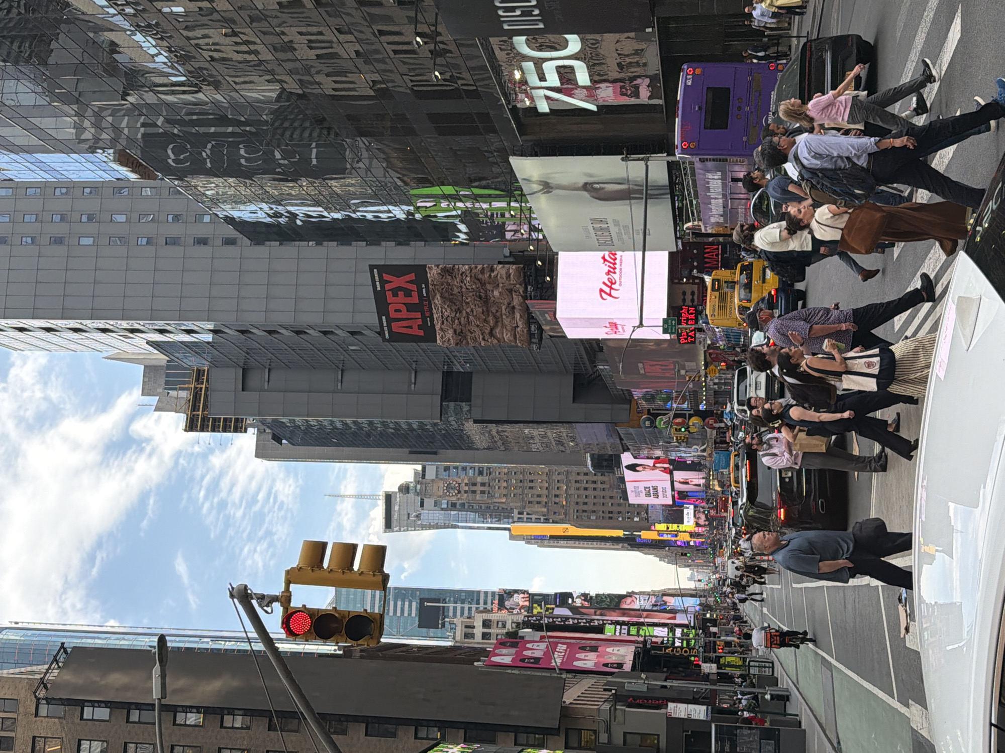 The image captures a vibrant scene in Times Square, New York City, USA, with a busy pedestrian crosswalk in the foreground. A crowd of people is seen crossing the street, with cars and buses navigating the traffic. Above the street, a multitude of billboards and digital displays adorn the buildings, showcasing advertisements and various messages. A traffic light is visible, displaying a red light, indicating that pedestrians should not cross at that moment. The architecture is typical of the area, with tall skyscrapers and a mix of older and modern buildings. The sky is partly cloudy, suggesting a daytime setting. The overall atmosphere is energetic and bustling, characteristic of Times Square.