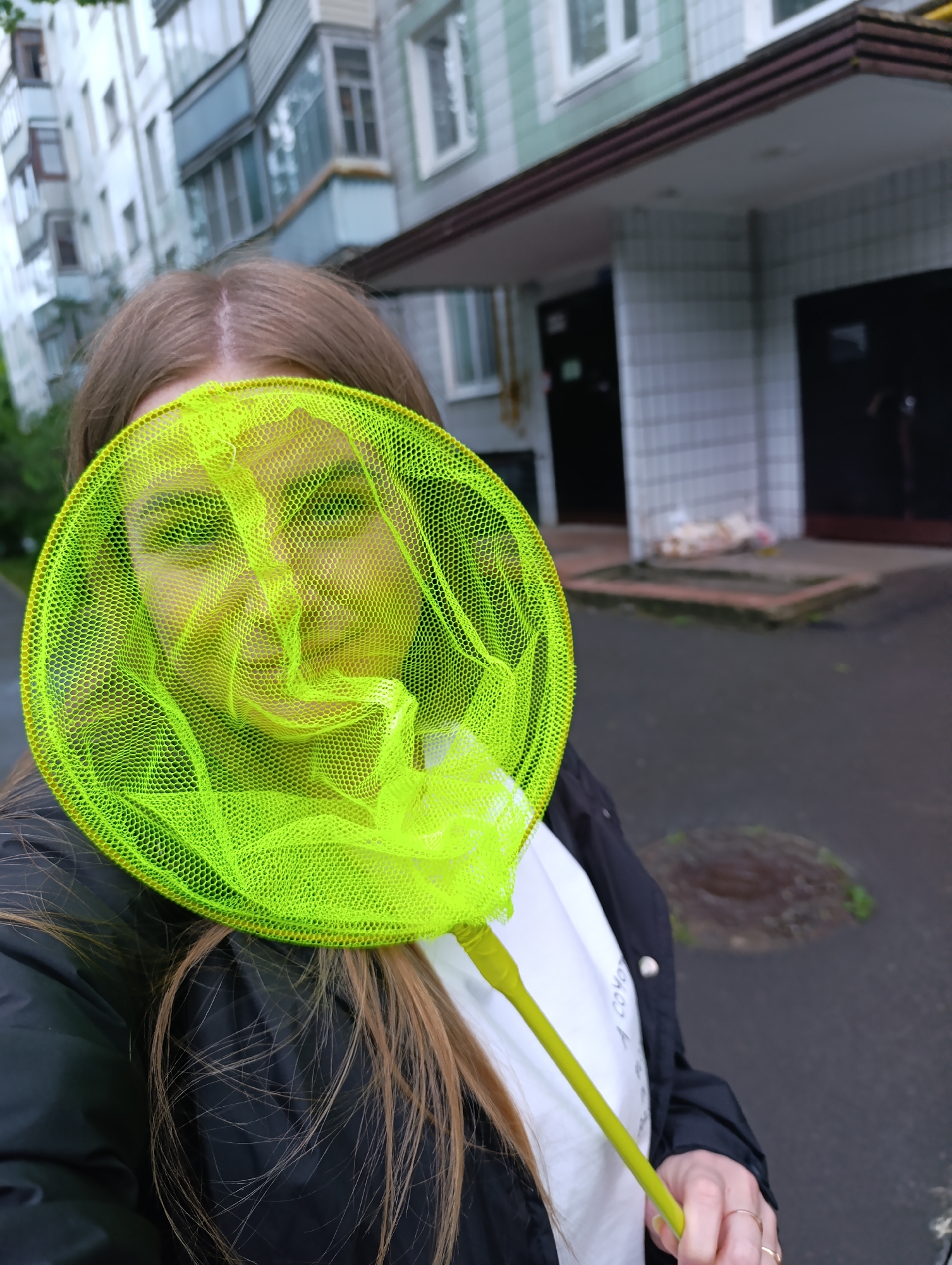 A medium shot depicts a woman holding a bright green circular net in front of her face. The net's mesh partially obscures her features, giving her eyes a green tint. She is wearing a black jacket over a white shirt. Her long, light brown hair is visible around the edges of the net.
The background features an apartment building with light gray and white tile-like exterior and dark doors. The ground appears to be wet asphalt. The location seems to be an urban area.