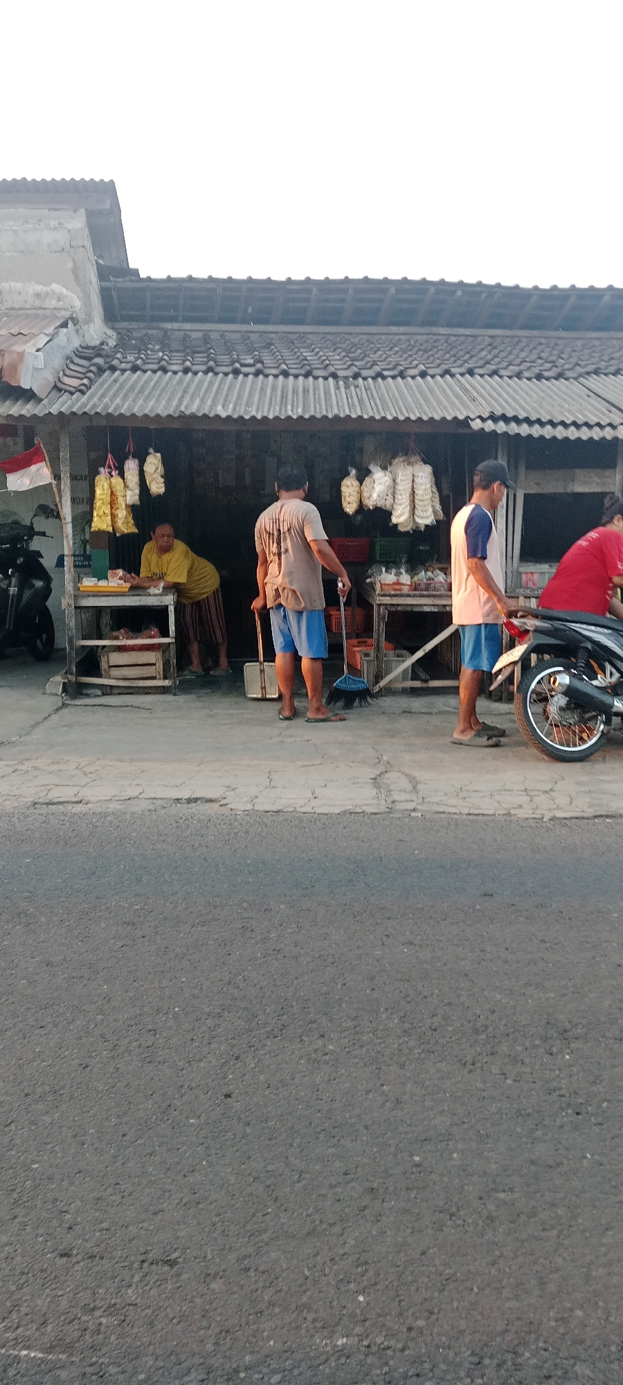 A street scene in Maguwoharjo, Indonesia, features a small storefront with several people and a motorcycle. The storefront has a corrugated metal roof and displays various goods, including bags of snacks. An elderly person in a yellow shirt and striped pants stands behind a counter. Another person wearing a brown t-shirt and blue shorts is sweeping the ground in front of the store with a broom and dustpan. A third person, wearing a light pink shirt with blue trim and blue shorts, stands near a motorcycle. A fourth person in a red shirt is partially visible near the motorcycle. The ground in front of the storefront is paved, and a road is visible in the foreground. The sky is bright and appears to be early morning or late afternoon.