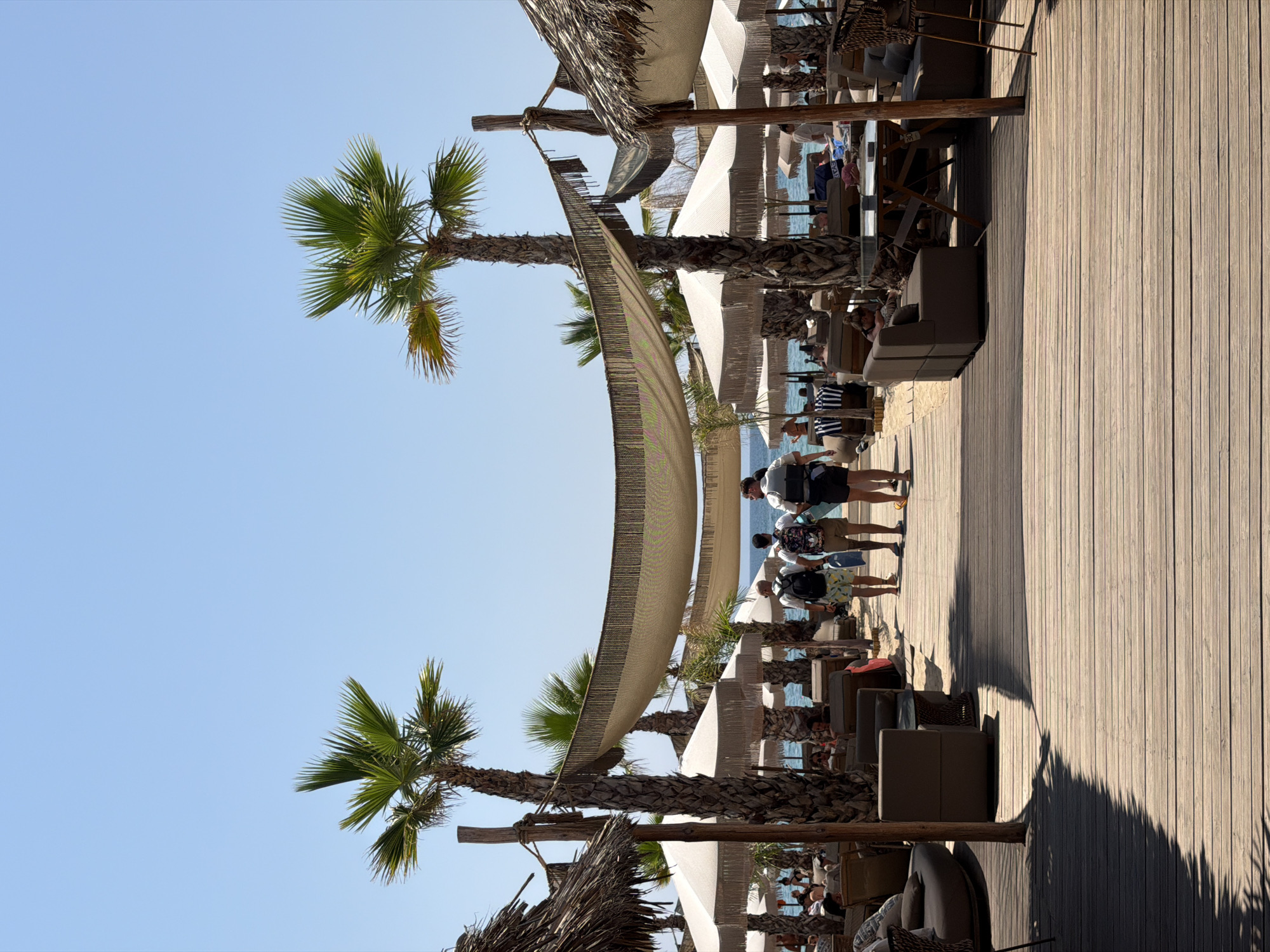 The image shows a view down a wooden boardwalk at a beachside establishment in Ofrynio Beach, Greece. The boardwalk is lined with palm trees, thatched umbrellas, and lounge seating. Several people are walking away from the camera down the boardwalk, and others are seated at tables or lounging on chairs. The sea and a clear blue sky are visible in the background. The scene suggests a relaxed, sunny day at a beach resort.