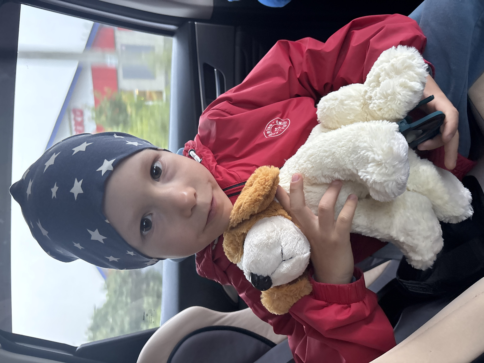 A young child is depicted inside a car, seated in what appears to be a car or booster seat. The image is rotated 90 degrees clockwise. The child, likely a boy, has dark eyes and is wearing a dark blue cap adorned with white stars, along with a bright red jacket. On the left chest area of his jacket, a circular white logo is visible, featuring partially legible text that appears to be "BRIND" or "BRAND" and "ON..." or "ORIGINAL".
The child is holding two stuffed animals: a large, fluffy white one to which a dark green clip is attached, and a smaller brown and white stuffed dog. He looks directly at the camera with a calm, neutral expression.
Through the car window behind him, an outdoor scene in Chaikovsky, Russia, is visible. This includes a building with a prominent red and white facade, featuring some blurry white lettering, possibly "OCO" or "OKO", on the red section. Green trees or bushes are also seen beyond the building. The sky appears overcast, indicating it is daytime. The scene captures a moment of travel.