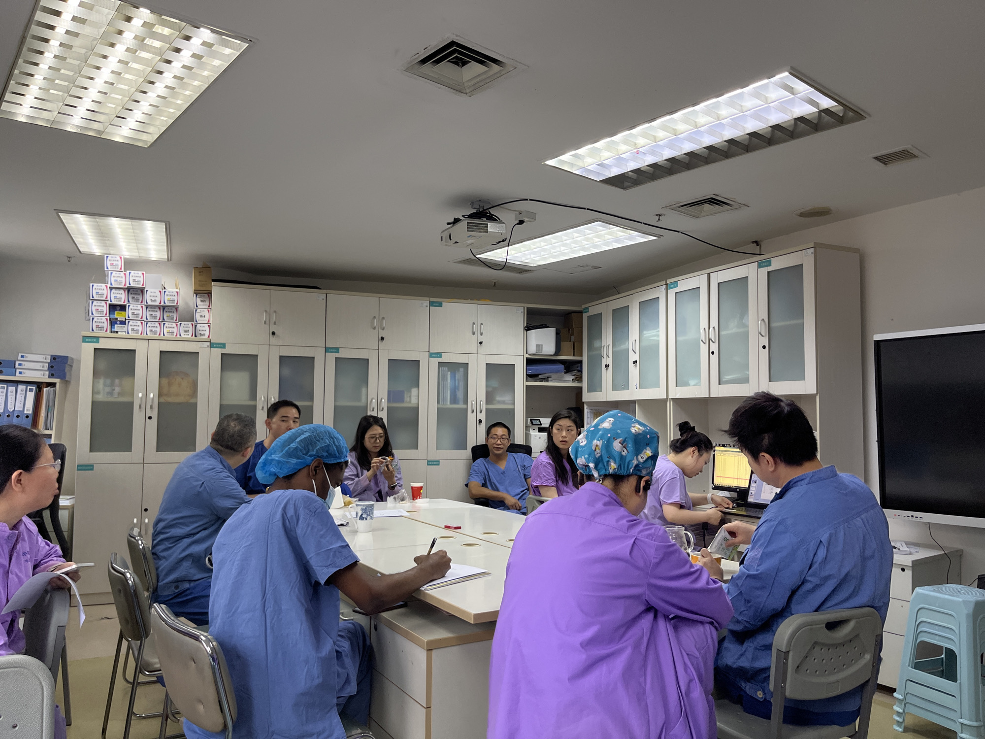 Location: A conference room in Yuzhong District, China.
Scene: A group of approximately ten people, likely medical professionals, are seated around a large table. Most are wearing surgical scrubs and head coverings. Some are writing on papers, while others are looking at laptops or interacting with each other.
Environment: The room is brightly lit with overhead fluorescent lights. Along one wall are numerous white cabinets with glass doors. A projector hangs from the ceiling. A stack of blue plastic chairs is visible in the corner.