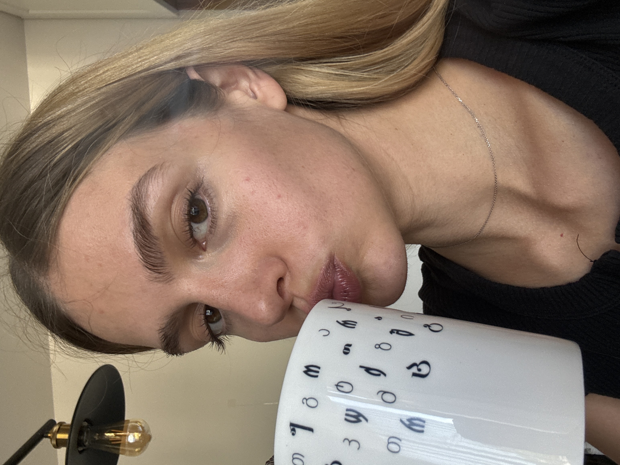 A woman with light skin, brown eyes, and blonde hair is holding a white mug with black numbers and symbols on it. She is wearing a black top and a thin silver necklace. In the bottom left corner of the image, there is a black desk lamp with a visible lightbulb. The location and city are unknown.