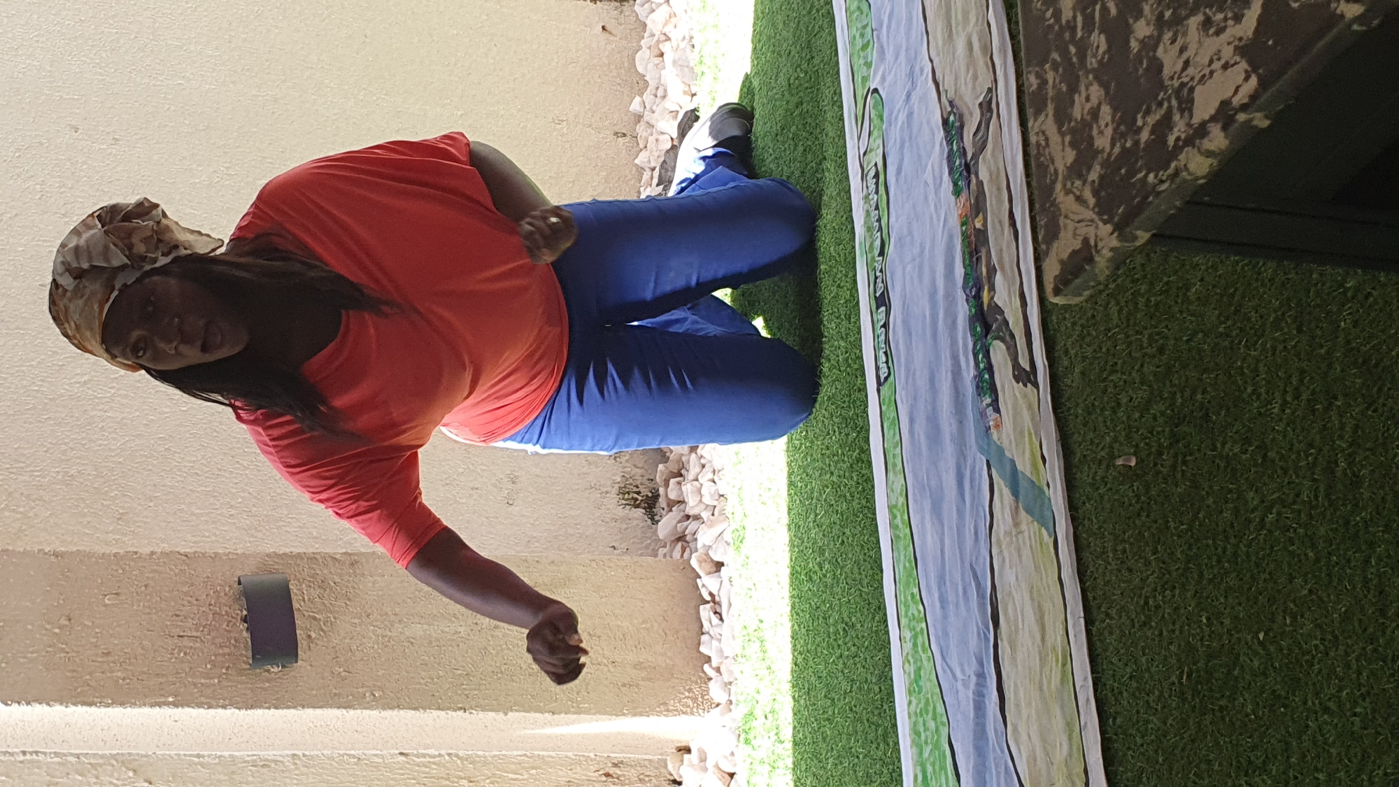 A Black woman, wearing a coral t-shirt, blue pants, and a patterned headscarf, kneels on artificial turf. She is facing forward and appears to be speaking animatedly, with her mouth open and her right arm extended. In the foreground, a large, rolled-up banner is visible on the turf. The background consists of a textured beige wall and a scattering of light-colored stones. The scene is outdoors, suggested by the natural lighting and the presence of turf. The location is identified as Gura Top, Nigeria.