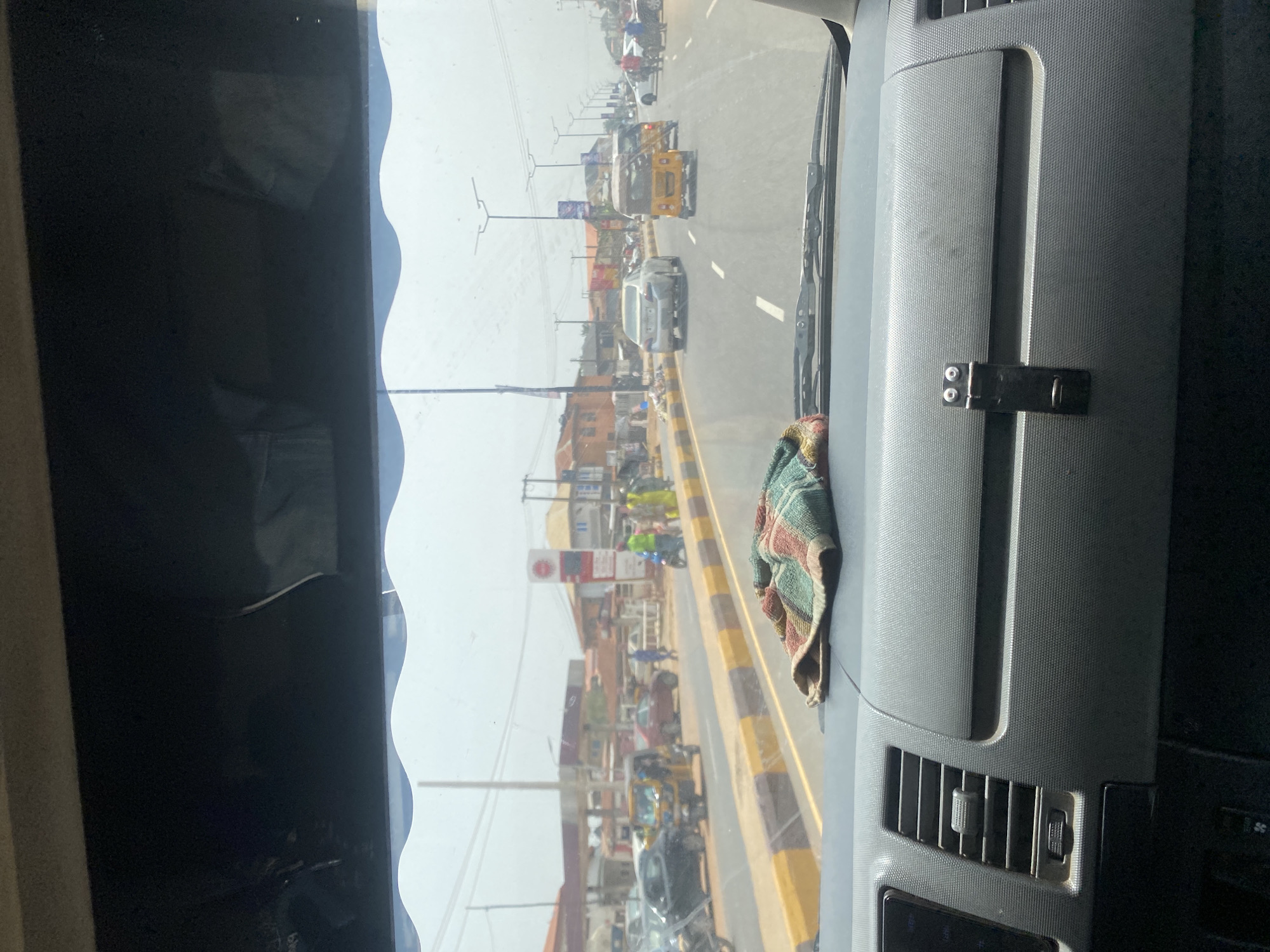 View from inside a vehicle traveling along a city street in Alakia, Nigeria. A light-colored sedan and a yellow tricycle taxi are visible, among other vehicles. People are walking alongside the road. Buildings and utility poles line the street. A fuel station is present. The dashboard of the vehicle is visible.