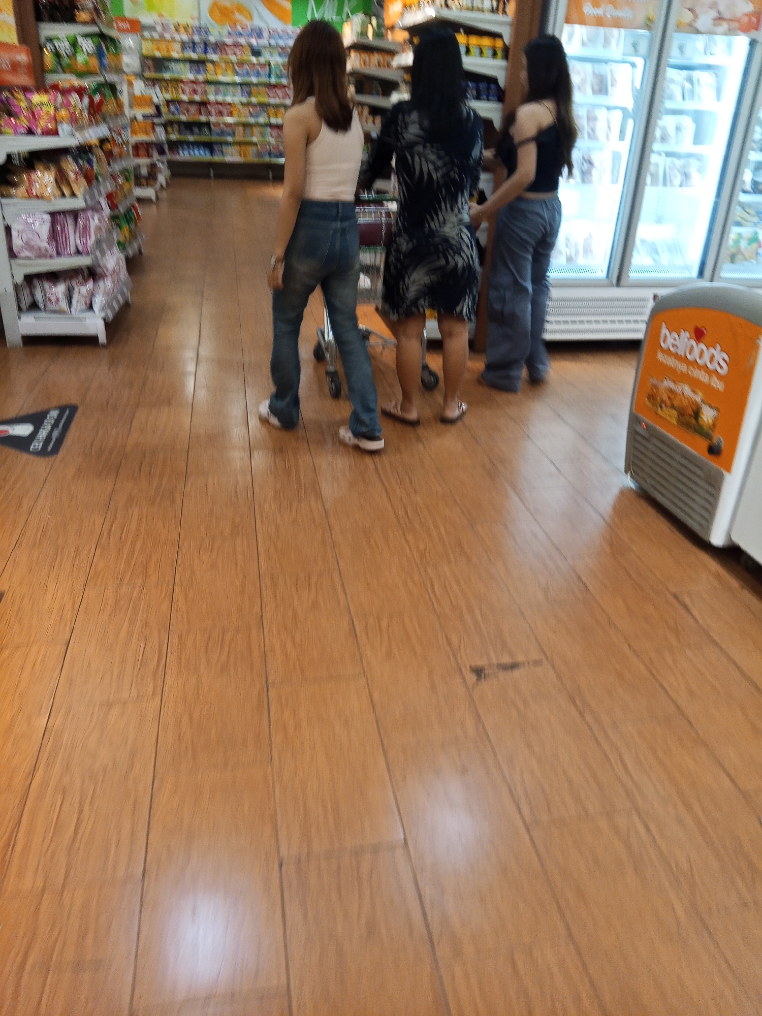 Three individuals are observed from the posterior in a supermarket aisle. The person on the left, wearing a pink tank top and blue jeans, stands with their back to the viewer, facing away from the camera. To their right, a person in a black and white patterned dress is also facing away. The individual on the far right, wearing a black tank top and grey pants, is also seen from behind, facing the same direction as the other two. They are positioned near a refrigerated display case with a "bellfoods" logo visible on an orange sign. The floor is constructed of polished wooden planks. Shelving stocked with various products lines the left side of the image, indicating a retail environment. The overall scene depicts a casual shopping excursion within a commercial establishment. The presence of the "bellfoods" logo suggests a potential connection to a food vendor or distributor, and the general store layout is consistent with a supermarket in Lubuklinggau, Indonesia.