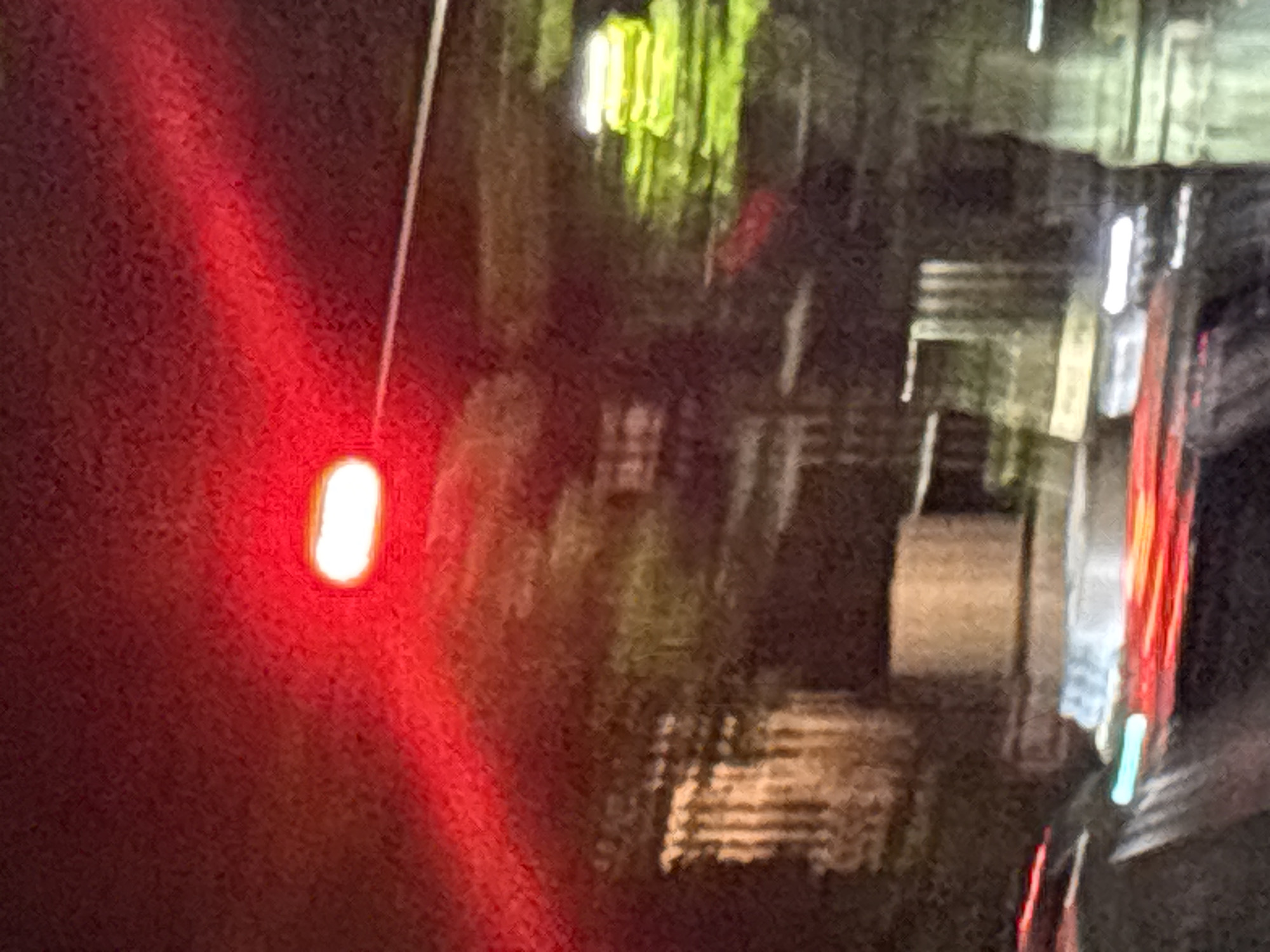 The image depicts an urban scene at night, possibly in Buenos Aires, Argentina, given the street and building styles. A blurred, bright red light source is prominent on the left, appearing to be a taillight or signal. The background shows indistinct building facades with illuminated windows and vertical lighting features. The overall image is blurred and lacks sharpness, indicative of low-light conditions and possible motion.