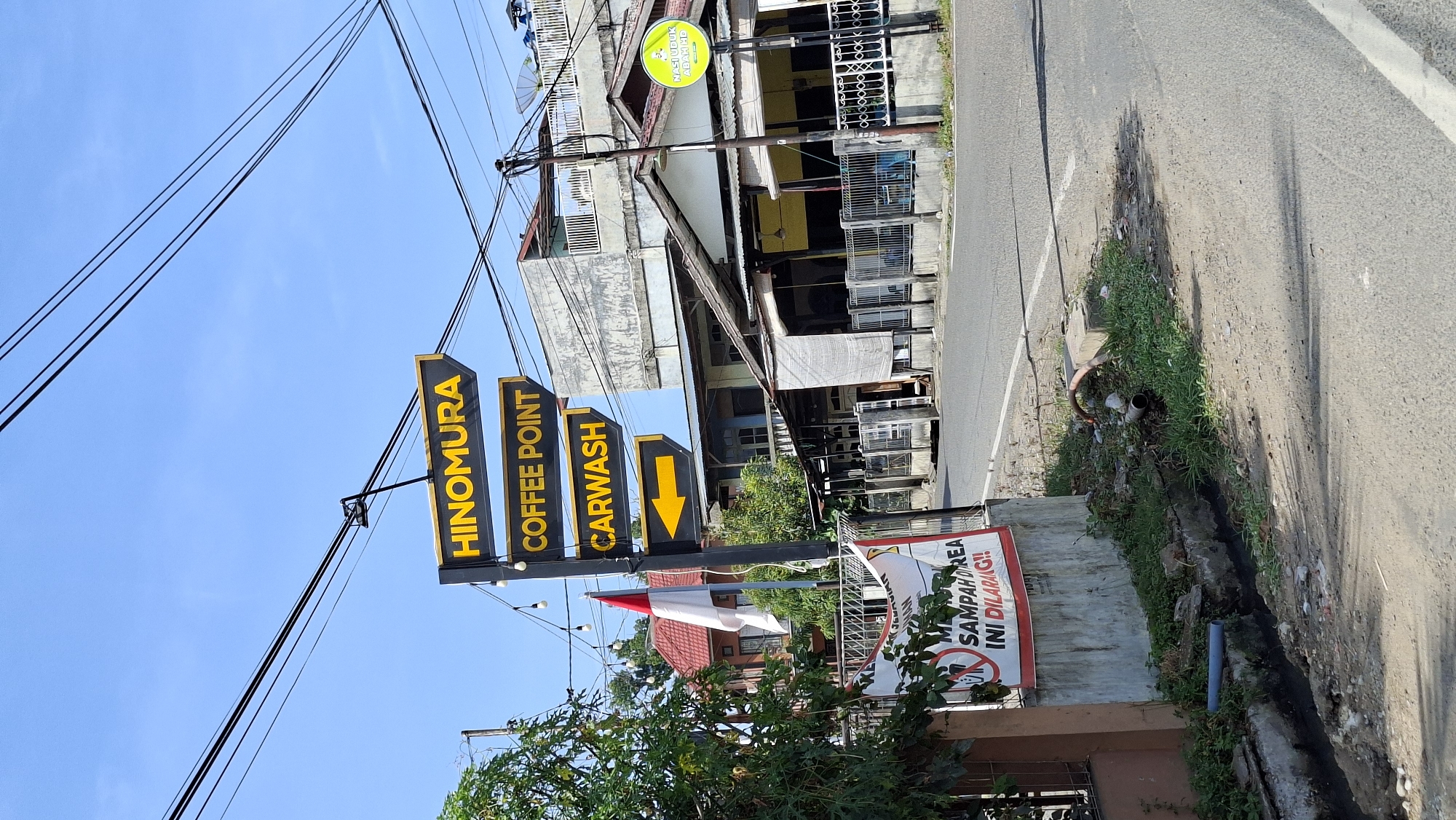 The image depicts a street scene in Lhokseumawe, Indonesia, with a clear blue sky overhead and power lines crisscrossing the frame. A prominent directional sign with yellow lettering on black backgrounds points to "HINOMURA," "COFFEE POINT," and "CARWASH," with a yellow arrow indicating the direction of "CARWASH." To the left of the sign, an Indonesian flag hangs on a flagpole. In the foreground, a small concrete barrier and a drainage ditch are visible, with some debris and vegetation. Beyond the sign, buildings line the street, including what appears to be a restaurant or shop with outdoor seating. Another sign with a yellow circular logo and the text "NASI UDUK ABAH HI" is also present. The overall scene suggests a commercial area with various services available.