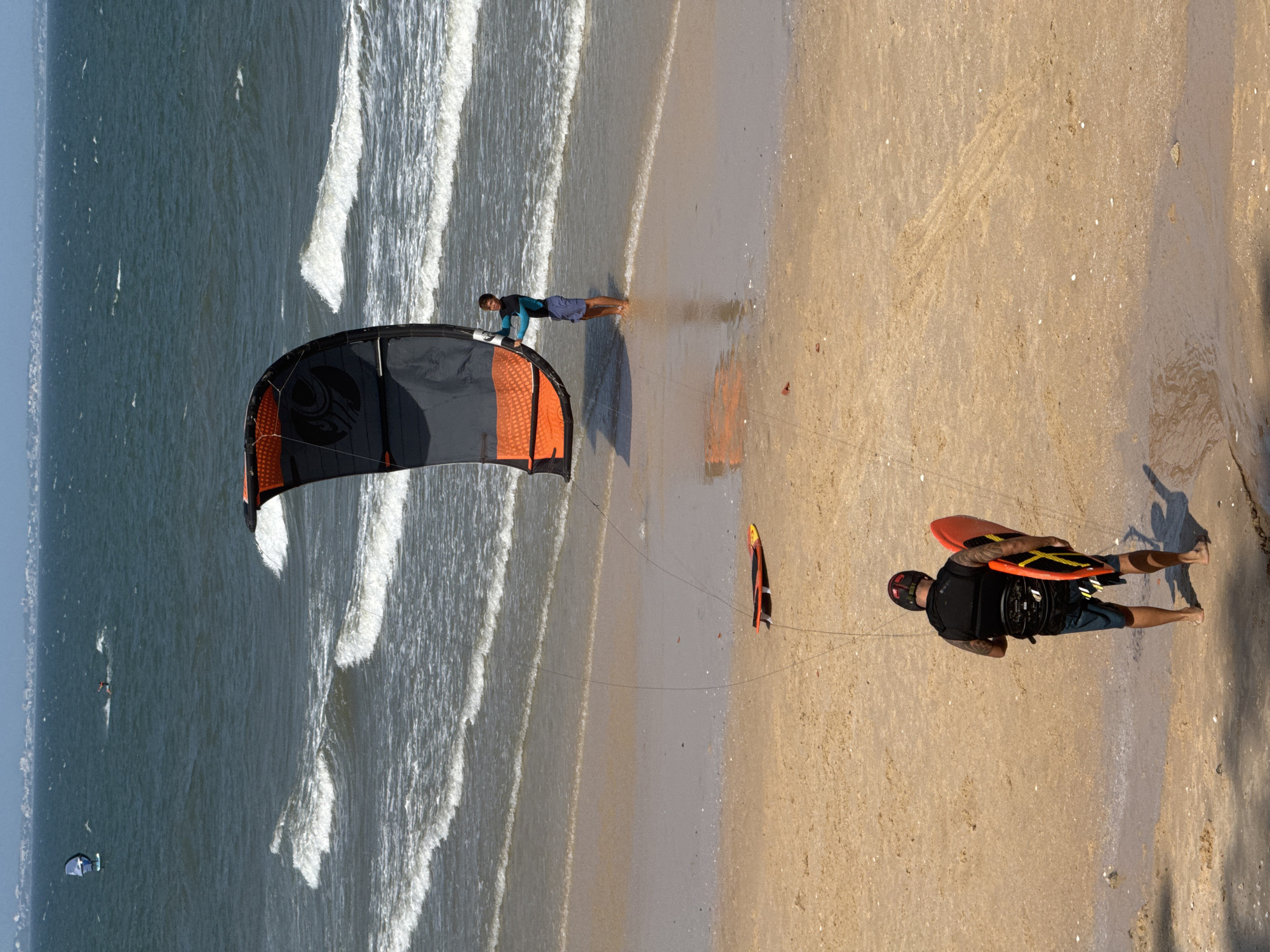 In Phan Thiết, Vietnam, on a sandy beach bordering the ocean, a kitesurfer stands waist-deep in the water, holding a large, dark grey and orange kite. His body is positioned to control the kite, which is partially inflated. The water is a deep blue-green, with white-capped waves rolling towards the shore. Further out, another person is kitesurfing, a small figure on the water. Closer to the shore, a second kitesurfer, wearing a black vest and dark shorts, stands on the wet sand, holding a bright orange board. The beach is sandy and shows signs of being recently wet, with some scattered shells and debris. The sky is a pale blue, suggesting a sunny day. The scene captures a moment of recreational activity on the coast.