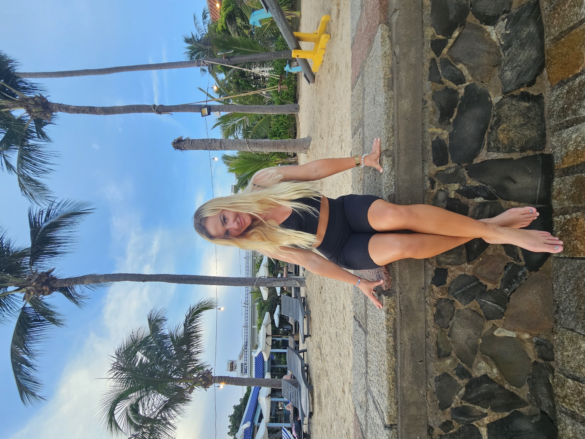 A blonde woman is sitting on a stone wall at the beach in Phu Thuy Ward, Vietnam. She is wearing a black sports bra and black shorts. Her legs are crossed, and her feet are bare. Behind her, palm trees, lounge chairs, and umbrellas are visible on the sand. In the background, there is a playground with a seesaw and a swing. The sky is cloudy and blue. The lighting suggests it is either early morning or late afternoon.