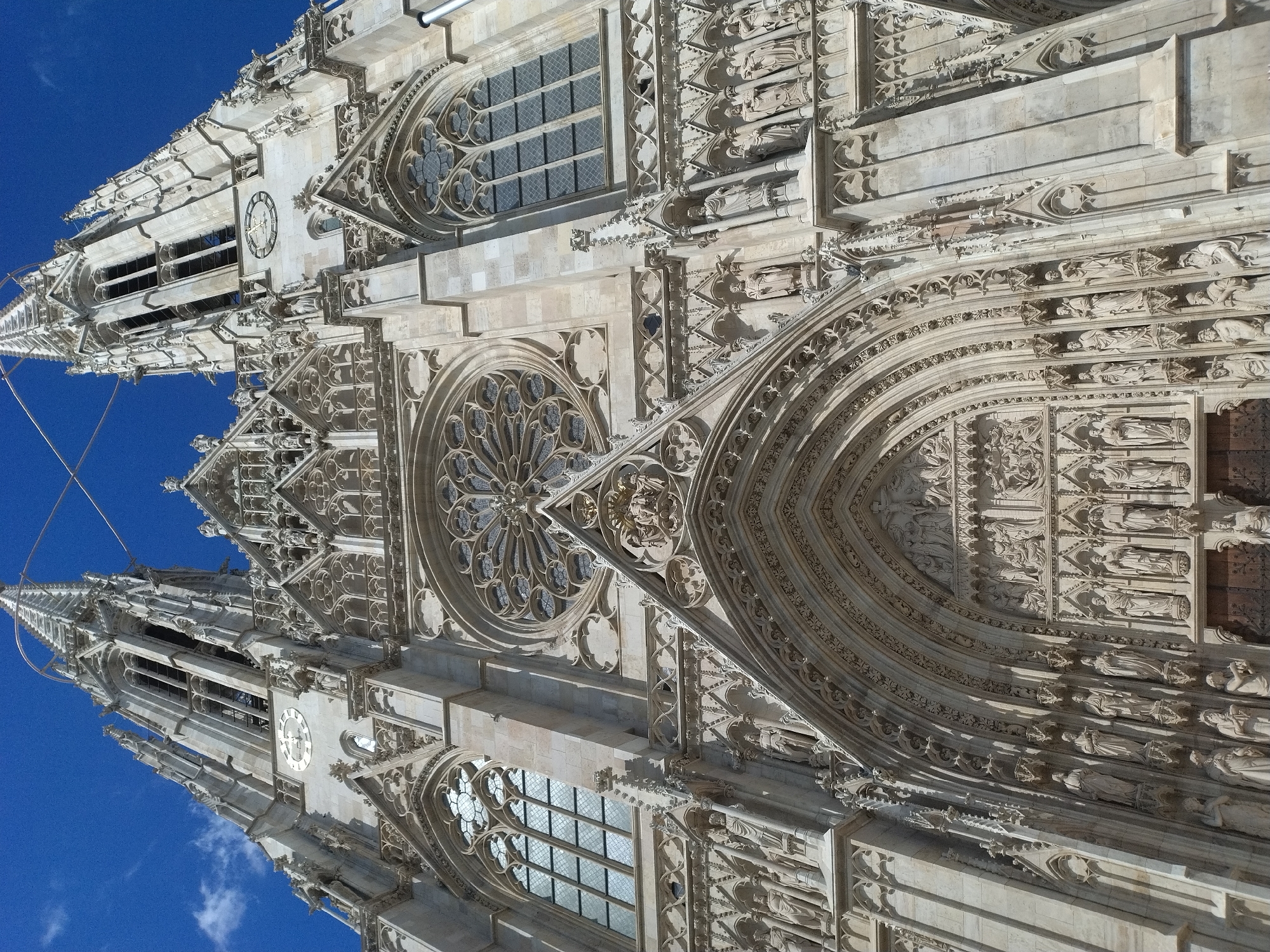 The image shows a low-angle, full shot of the facade of St. Stephen's Cathedral (Stephansdom) in Vienna, Austria, under a clear blue sky with a few scattered clouds. The cathedral is constructed of light-colored stone and features intricate Gothic architecture. Dominating the frame are the two prominent spires, each topped with a sharp, pointed roof. Below the spires, the facade is adorned with numerous detailed sculptures and carvings, including statues of figures in niches, a large rose window, and decorative tracery. The main entrance is a grand arched portal, also heavily ornamented with religious figures and scenes. The detail suggests the facade is well-maintained. The perspective emphasizes the verticality and grandeur of the cathedral.