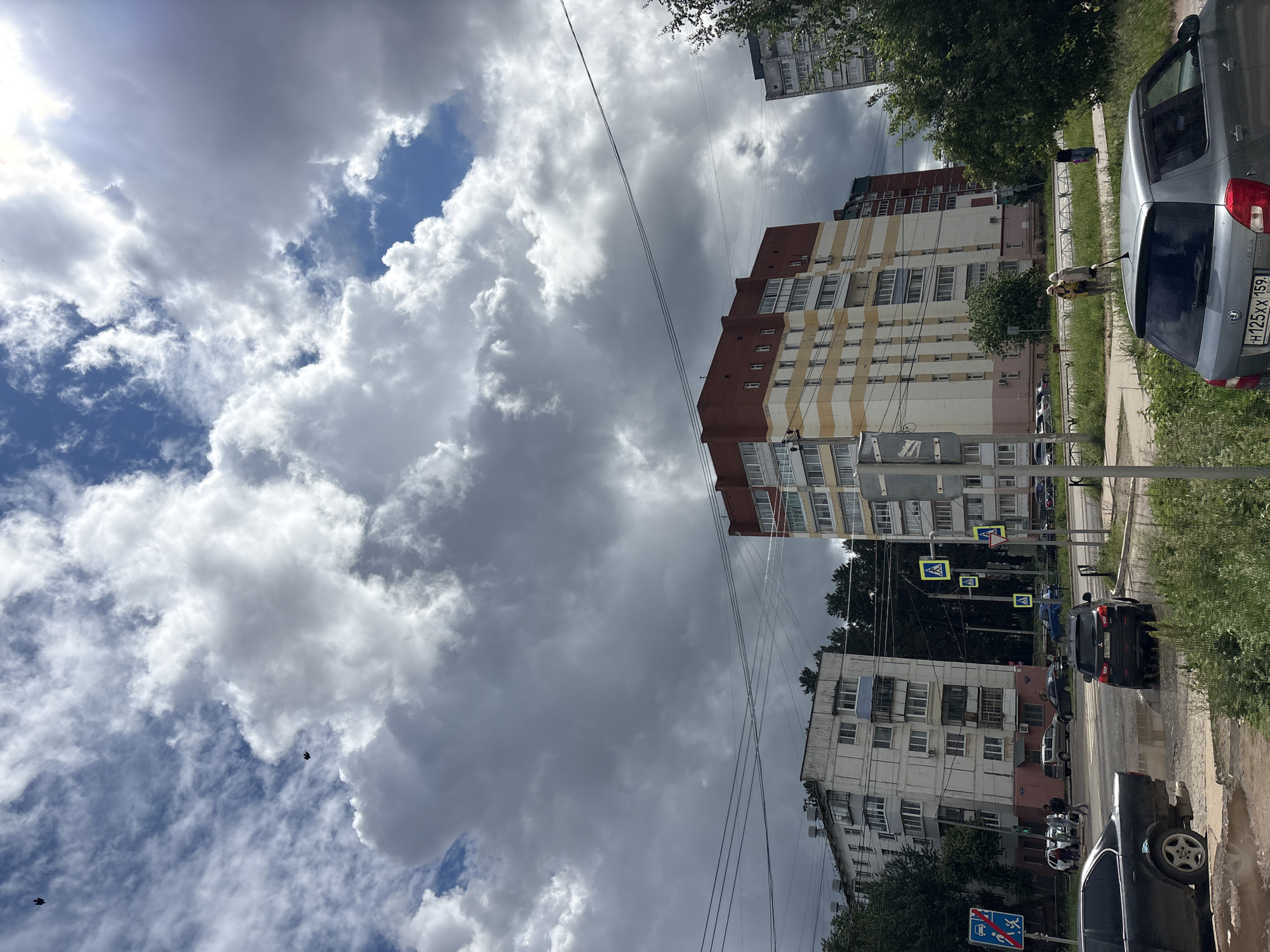 This outdoor shot, taken in Chaikovsky, Russia, captures a residential street scene under a dynamic, partly cloudy sky. Large, dramatic cumulus clouds dominate the upper portion of the frame, interspersed with patches of clear blue sky and bright sunlight breaking through on the left.
The scene features several multi-story apartment buildings. A prominent building on the right has a distinctive red roof and a facade painted with alternating light yellow and white stripes. Below it, an older, greyish-white building with multiple balconies and air conditioning units is visible. Power lines crisscross the sky between the buildings.
At street level, a road runs through the center-right of the image, flanked by sidewalks and grassy areas with trees and bushes. Several cars are present; a silver-grey car with the Russian license plate "H125XX 159" is prominently parked on the right, partially obscured by foliage. Further down the street, other vehicles, including a black car, are parked or driving. Two people are visible near the grey car, one appearing to hold a long stick or pole. Another individual is seen walking on the sidewalk in the distance.
Visible street signage includes multiple yellow square signs indicating a pedestrian crossing, and a blue rectangular sign with a white figure walking with a red diagonal line, suggesting a restriction, possibly "no pedestrian access" or similar, below which is a smaller blue sign that might denote a public transport stop. The time of day appears to be daytime, with ample natural light despite the heavy cloud cover. The overall impression is a typical urban residential environment.