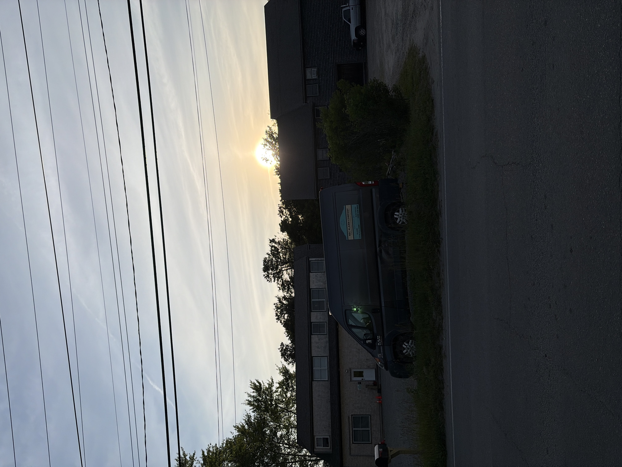 An outdoor scene is depicted during late afternoon or early evening, characterized by low sun angle, in Nantucket, United States of America. The foreground features an asphalt road with a continuous white line on its right edge, showing minor surface cracks. Adjacent to the road, a strip of unmaintained grass and weeds fronts a gravel lot.
In the midground, a dark-colored cargo van, likely a Ram ProMaster, is parked facing left on the gravel. Its side features a decal for "NEC ALARM COMPANY," displaying "ALARM (781) 834-3996" and "ELECTRICAL (508) 228-2859." A green light is illuminated on the van's driver's side. Behind the van and further back, two buildings are present. The left building is a two-story structure with grey shingle siding, multiple windows, and a white entry door. A small mailbox on a wooden post stands to its far left, and two orange traffic cones are positioned near its entrance. The right building is a darker, multi-story structure, possibly of log or dark shingle construction, with visible windows and a large, dark opening on its lower level. A dense green shrub partially obscures the view of the right building and a light-colored pickup truck parked beyond it in the gravel lot. The sun, a bright yellow-orange orb, is low in the sky, positioned above and behind the right building and partially obscured by trees.
The background consists of various trees, primarily coniferous, silhouetted against the sky. The sky transitions from a light blue at the top to a pale yellow closer to the horizon. Approximately ten dark utility lines traverse horizontally across the upper portion of the sky, with a faint, diagonal contrail visible in the upper-left quadrant. No people are present in the image.