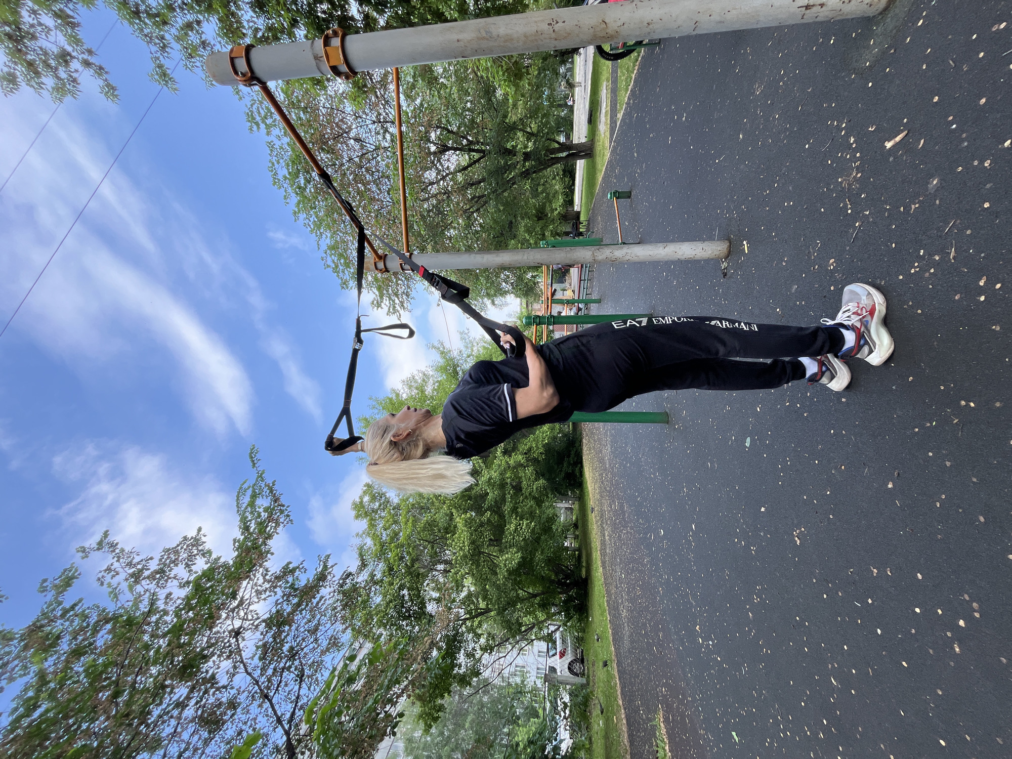 A blonde woman in black workout attire is exercising using suspension trainers on an outdoor sports ground. She is leaning back, holding onto the handles of the trainers. The sports ground has a black rubberized surface with scattered leaves. In the background, there are trees, a multi-story building, parked cars, and other exercise equipment. The sky is blue with scattered white clouds. The scene is consistent with an outdoor fitness area in a residential neighborhood, likely in Krasnoyarsk, Russia, given the architectural style of the building and the general environment.