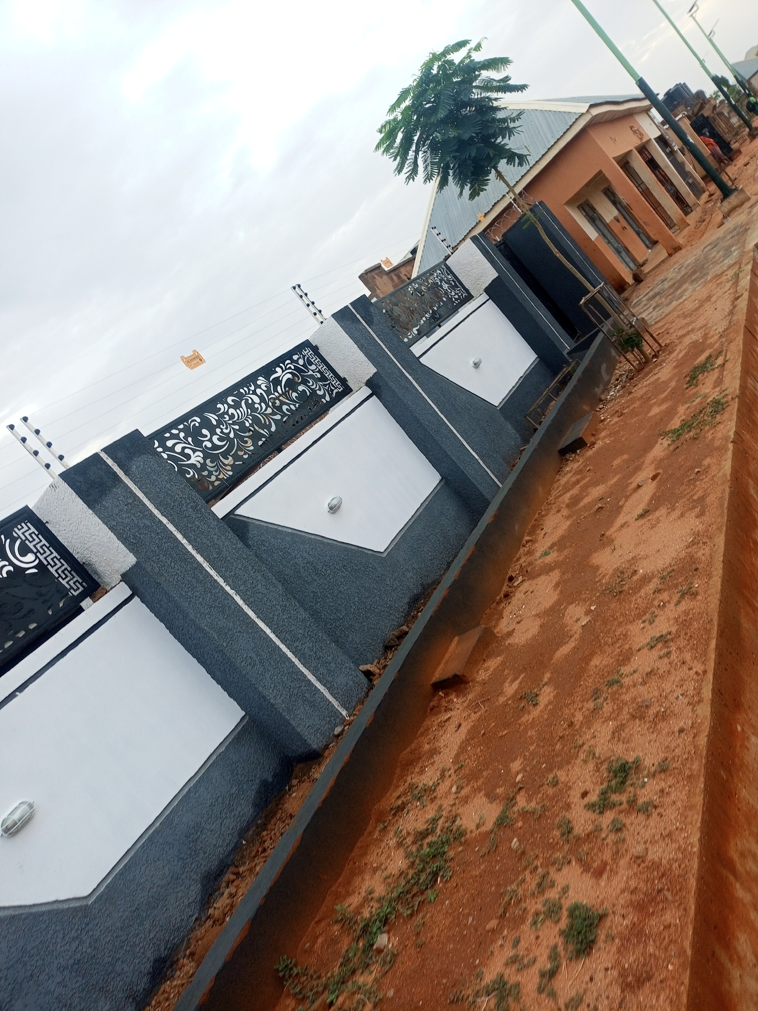 The image shows a residential fence in Tempure, Nigeria. The fence has a gray, textured base with white panels and decorative black metalwork on top. There is an electric fence running along the top. The ground is reddish-brown dirt and grass. A gutter runs along the base of the fence. In the background are houses and streetlights. The sky is overcast.