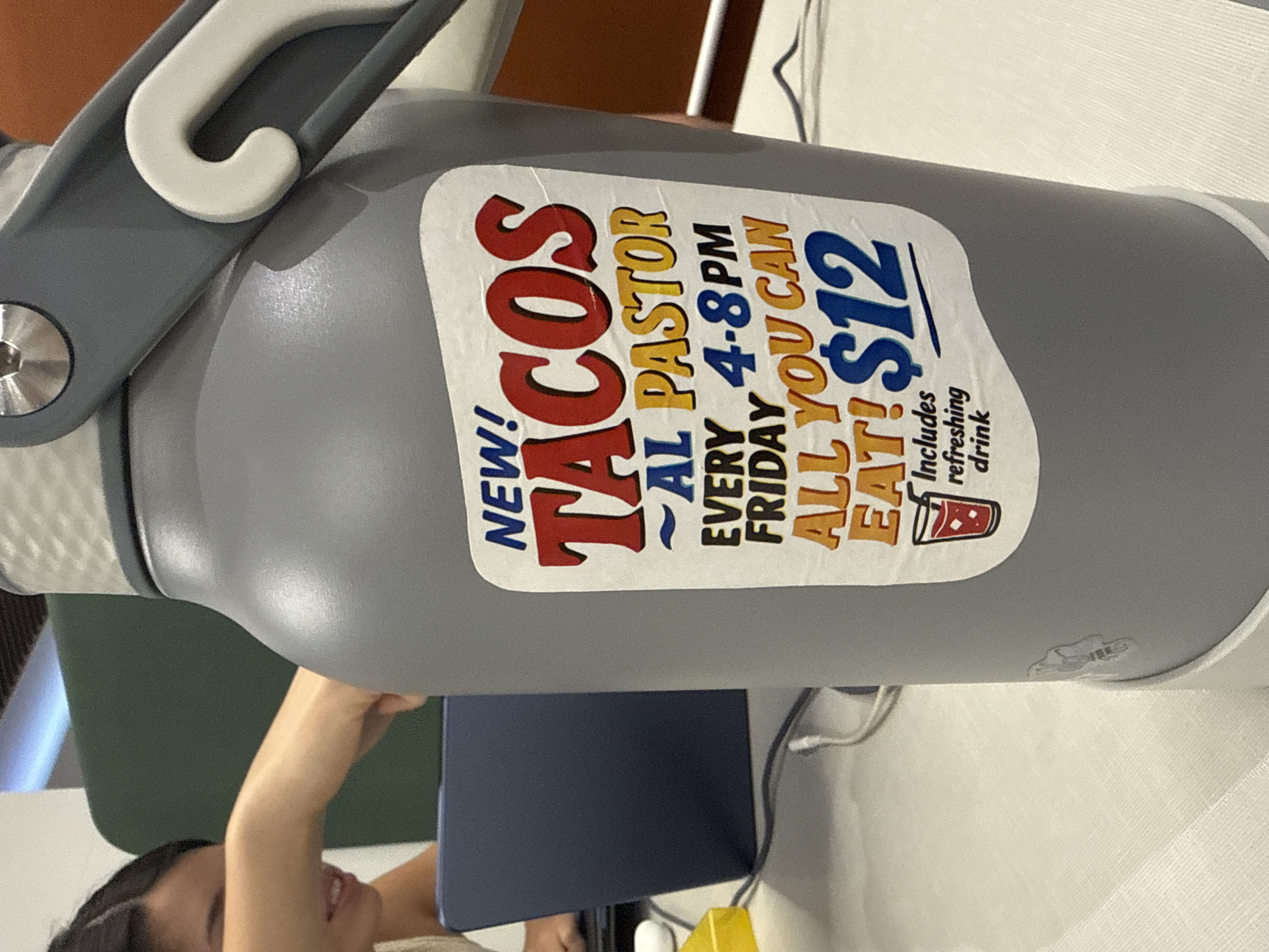 A gray, insulated bottle, possibly a water bottle or thermos, is prominently featured in this close-up image. Attached to its side is a white and gray handle. A white sticker adhered to the bottle advertises a food special. The text on the sticker reads: "NEW! TACOS AL PASTOR EVERY FRIDAY 4-8PM ALL YOU CAN EAT! $12 Includes refreshing drink." A small icon of a red cup with a straw is also visible next to the drink offer.
In the lower portion of the frame, a person is partially visible. Their bare arm, shoulder, and a smiling profile of their face suggest they are looking down or to the side. They appear to be holding or interacting with a dark blue electronic device, possibly a tablet or laptop. Several white cables and a yellow object, which could be a charger or small container, are also on the surface in front of them. The setting appears to be indoors, likely at a desk or table, with a light-colored surface dominating the background and hints of other furniture or walls. The image is oriented sideways. Given the context of "Al Pastor" tacos and the provided location, the advertisement likely pertains to a restaurant or food vendor in San Andrés Cholula, Mexico.