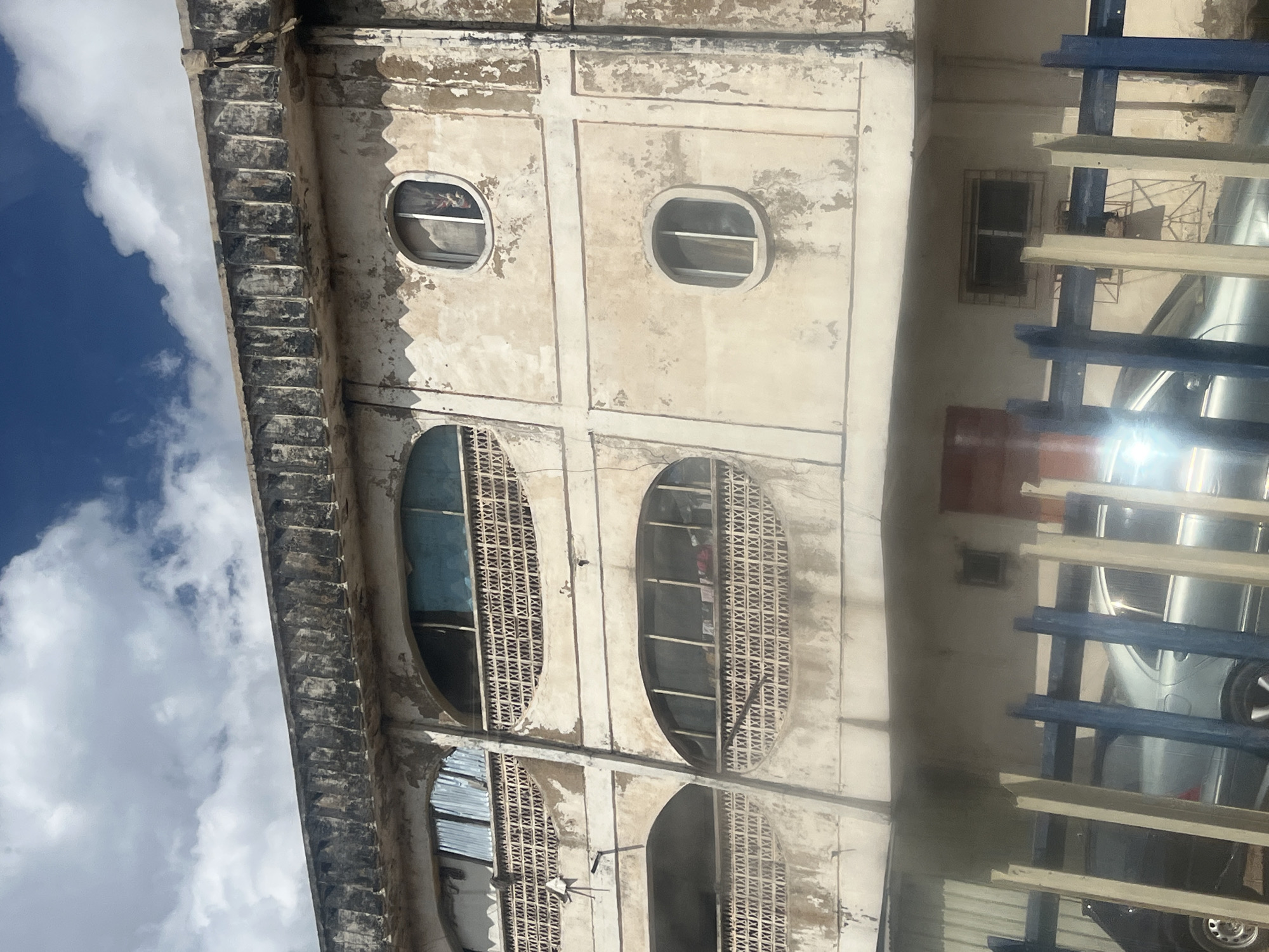 A close-up, eye-level shot captures the facade of a weathered, multi-story building in Jos, Nigeria, against a sky filled with scattered white clouds. The building's cream-colored walls are peeling and stained, revealing patches of what appears to be concrete underneath. The upper section of the building features a decorative, distressed concrete cornice with a series of vertical, ribbed elements.
Below the cornice, the second floor has two large, arched windows. The left window is partially obscured by corrugated metal sheeting, while the right window has a light blue curtain visible behind a latticework of crisscrossed patterns. A small, white object, possibly a satellite dish mount, is attached to the wall near the left arched window.
The third floor presents two more arched windows, similar in design to those above, but with different interior views. The left window reveals the same crisscrossed latticework, and the right window shows a light blue curtain.
Further down, the fourth floor features two oval-shaped windows. The left oval window is dark, with what appears to be a white curtain or blinds. The right oval window is also dark and shows a faint reflection. A second, similar oval window is visible on the floor below it.
At ground level, a blue and beige fence runs across the foreground, partially obscuring a silver sedan and a black vehicle parked behind it. The fence's wooden posts are arranged in a staggered pattern, with blue horizontal planks. A red door is visible behind the fence, set into a beige wall. The overall impression is one of age and disrepair, with signs of everyday life evident through the visible interiors of the building.