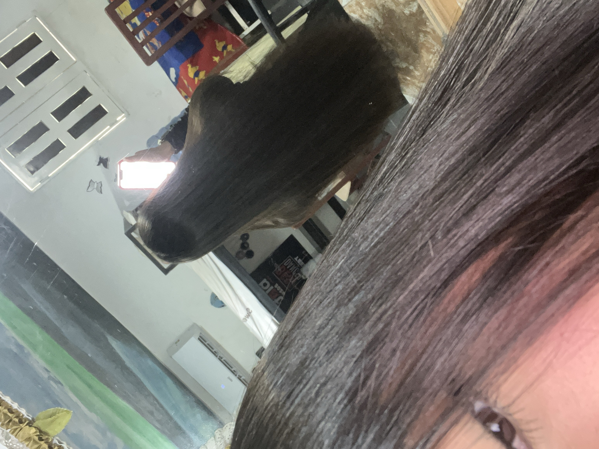 The image is a mirror selfie taken indoors in the city of Juan Griego, Venezuela. The lower-left foreground displays a partial view of a person's left eye with visible eyelashes and a light skin tone, alongside a mass of long, straight, dark brown hair flowing diagonally across the frame. In the central-right reflection, the back of a person with very long, straight, dark brown hair is visible. This individual holds a smartphone with a brightly illuminated, predominantly red-orange screen in their right hand, positioned to capture the reflection. A dark long-sleeved garment covers their right arm. The reflected background features light-colored walls. On the upper right, a white rectangular ceiling fixture or window frame with a grid pattern of four dark rectangular sections is present. The far-left wall reflects a multi-striped painting or mural displaying horizontal bands of green, blue, and dark grey/black, topped by a golden ruffled fabric decoration and a yellow leaf. Below this, a white wall-mounted air conditioning unit with horizontal vents and a blue indicator light is reflected. On the right, a dark brown wooden chair with vertical slats is visible, positioned near a surface covered with a colorful patterned fabric featuring red, blue, and yellow motifs. Two small, dark butterfly-shaped decals are affixed to the wall near the ceiling fixture, and a small blue object is visible near the air conditioner.