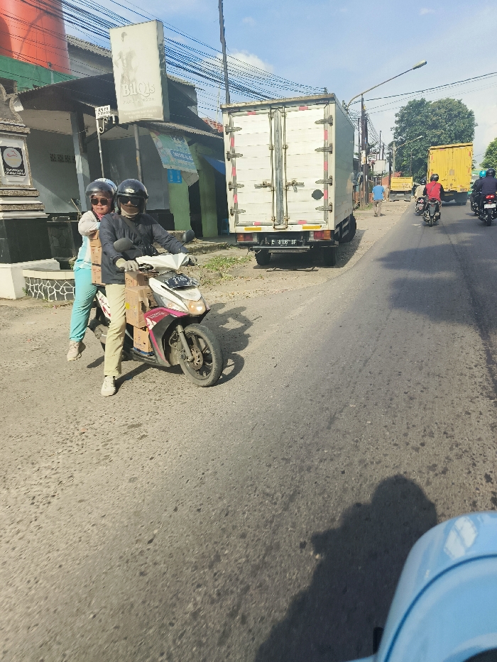 The image shows a man and a woman riding a motorcycle on a paved road. The man is wearing a helmet and is driving the motorcycle, while the woman is sitting behind him and wearing a helmet. The motorcycle is carrying several cardboard boxes. They are traveling along a two-lane road with a white truck stopped on the right. The truck is parked at the edge of the road with its rear doors closed. The woman is wearing a blue shirt and the man is wearing a dark shirt and khaki pants. Behind the motorcycle are several cars, including a yellow truck, and some people walking on the sidewalk. The scene is located in the city of Jatiluhur, Indonesia (latitude: -6.57, longitude: 107.438). The weather is sunny and the road is dry. The time of day is likely midday. There are buildings and trees in the background.