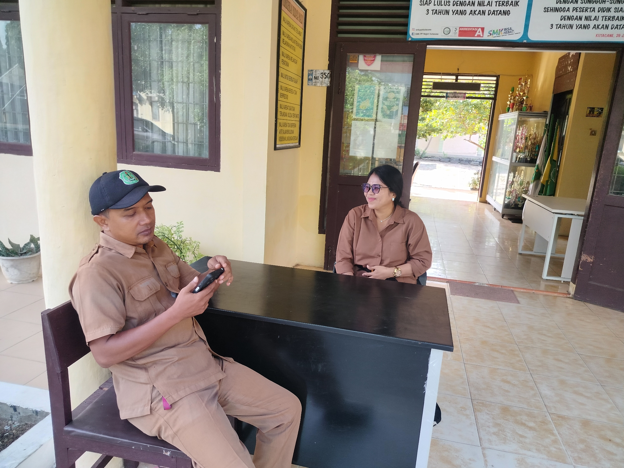 In Lawebekung, Indonesia, two individuals are seated near an entranceway to a building. On the left, a man wearing a brown uniform and a dark baseball cap sits on a dark wooden chair, holding a black smartphone. To his right, a woman in a brown button-down shirt and glasses is seated at a black desk, looking to her right. The environment includes a yellow-painted wall, a brown-framed window with sheer curtains, and a tiled floor. Behind the woman, a glass display case filled with trophies is visible, along with a doorway leading to an outdoor area with trees. A sign with Indonesian text is mounted on the yellow wall to the left of the man.